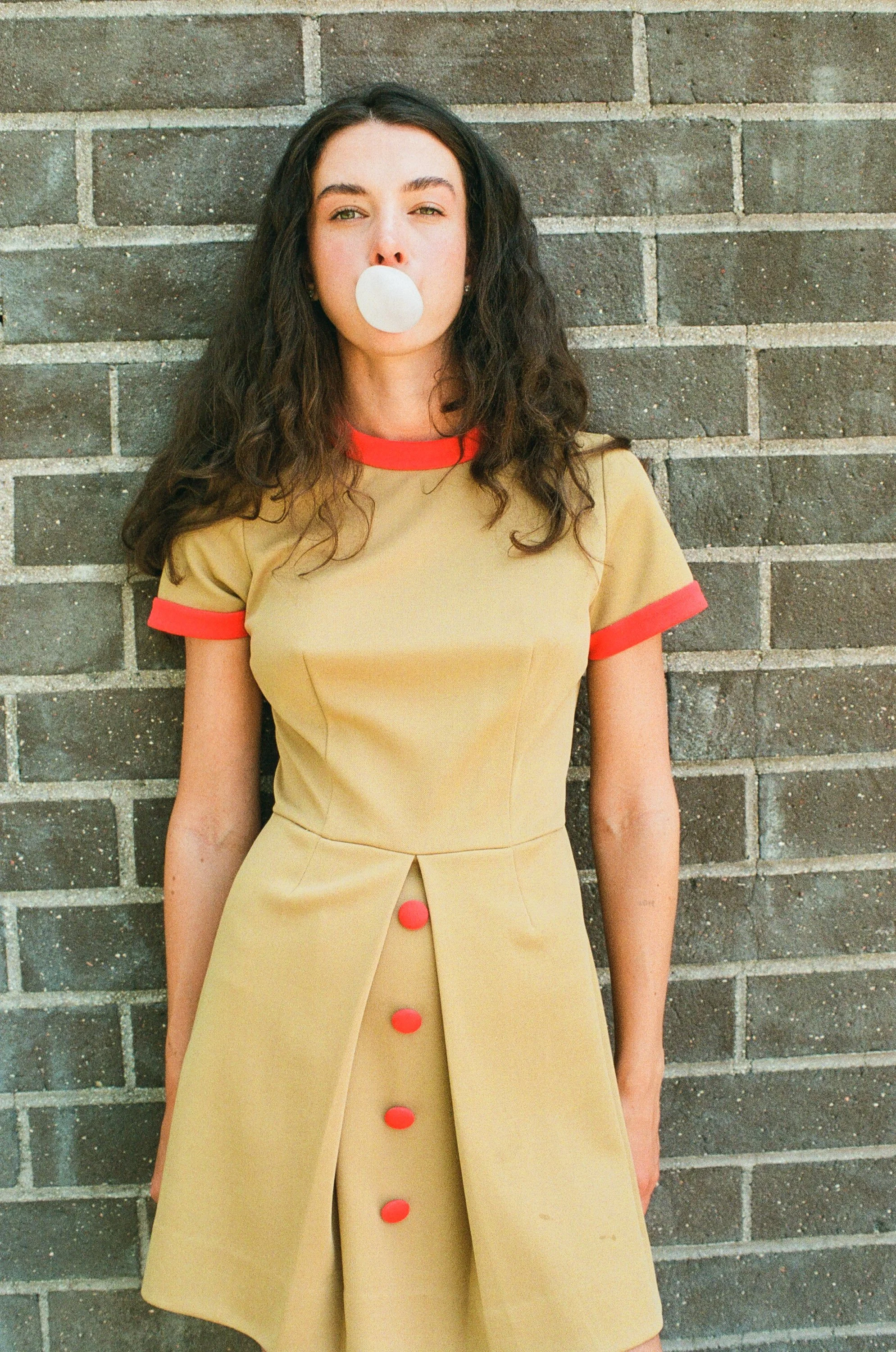 Model with long, dark wavy hair blowing a bubble gum bubble while wearing a beige dress with red accents against a brick wall.