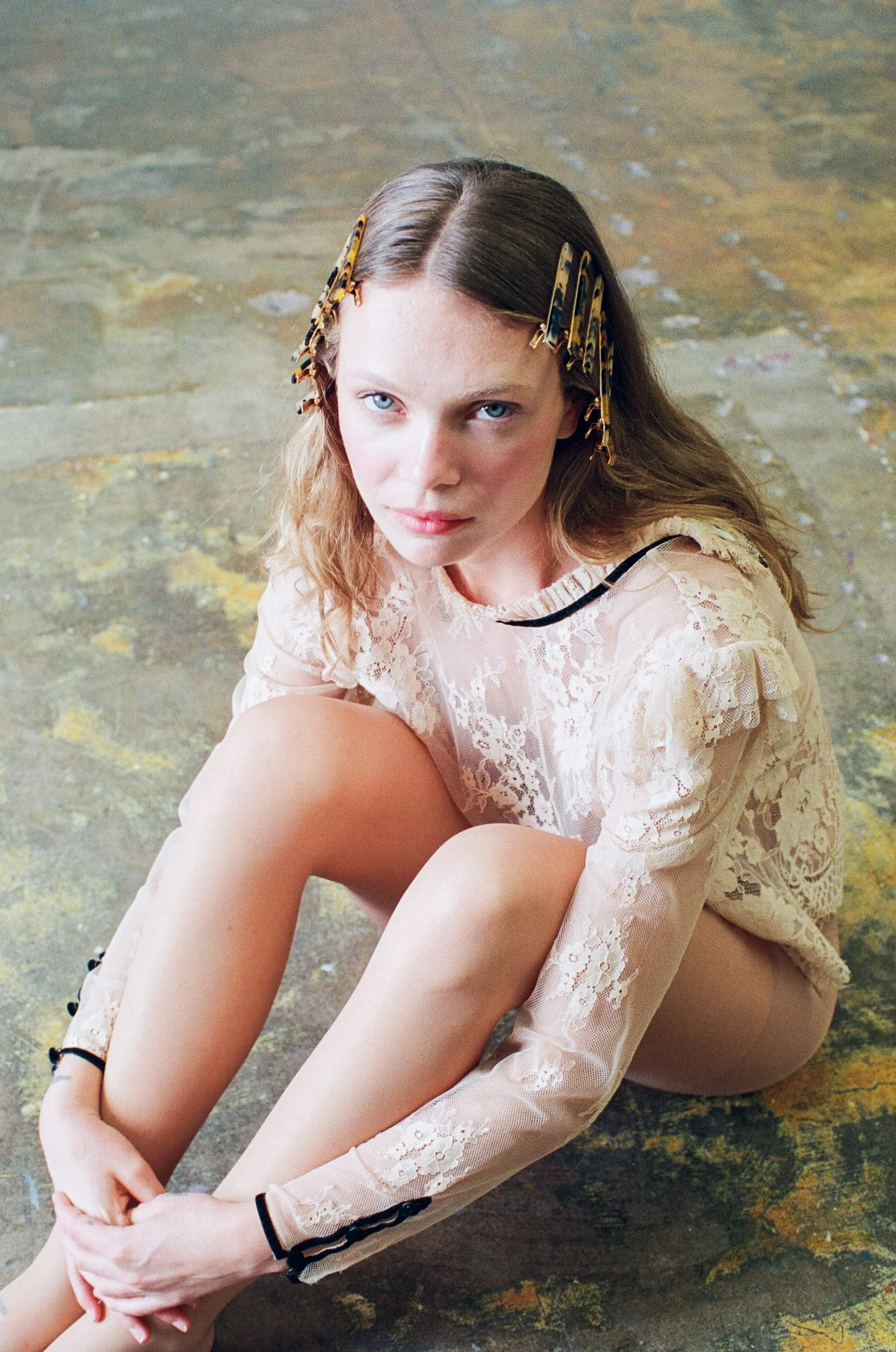 Fashion model with long light brown hair wearing a sheer white lace top with floral details and beige pants, sitting on a worn concrete floor with her arms wrapped around her legs while looking up at the camera.