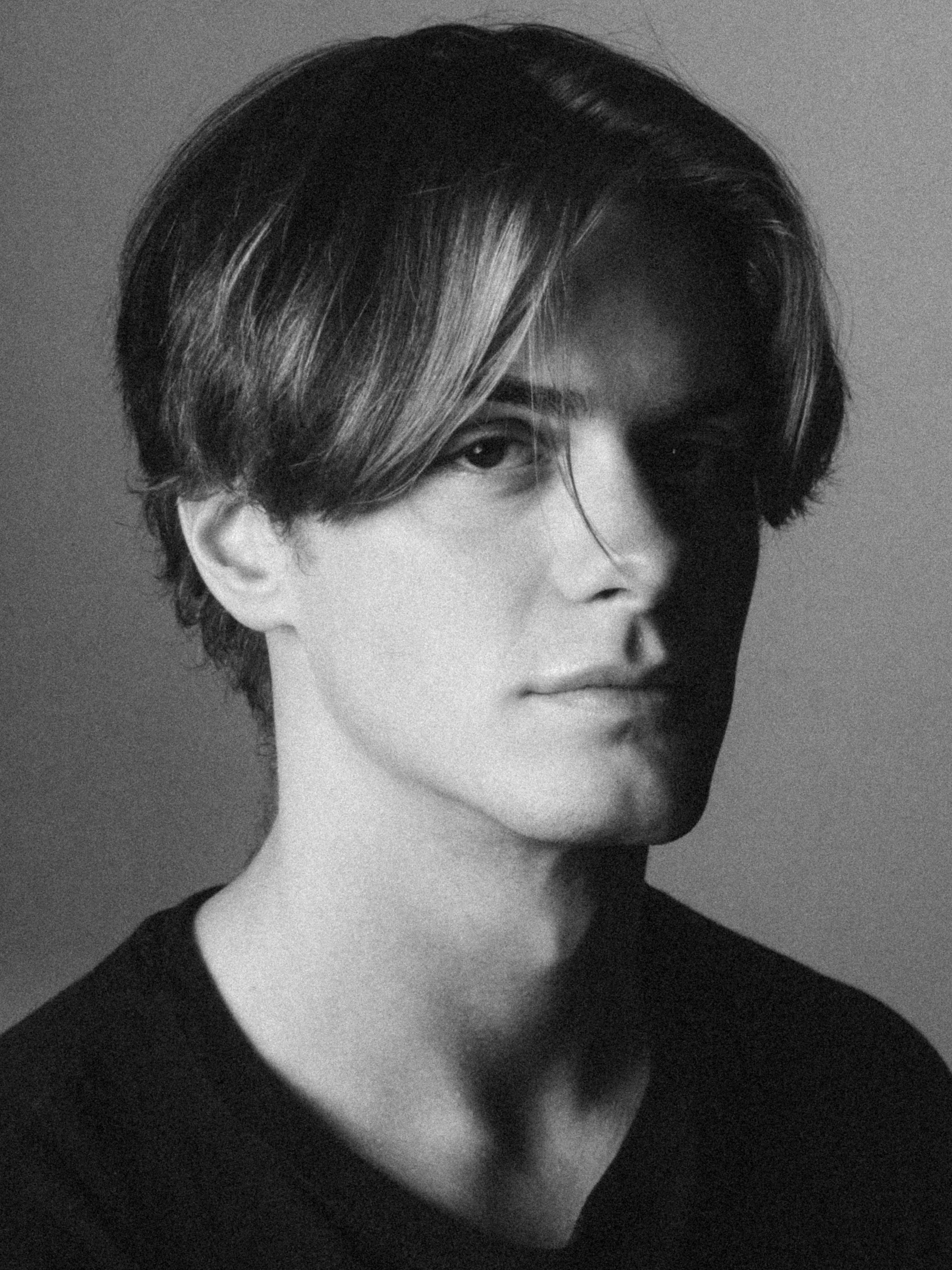 Black and white portrait of a person with short dark hair with a streak of lighter hair, wearing a dark top, looking slightly to the right.