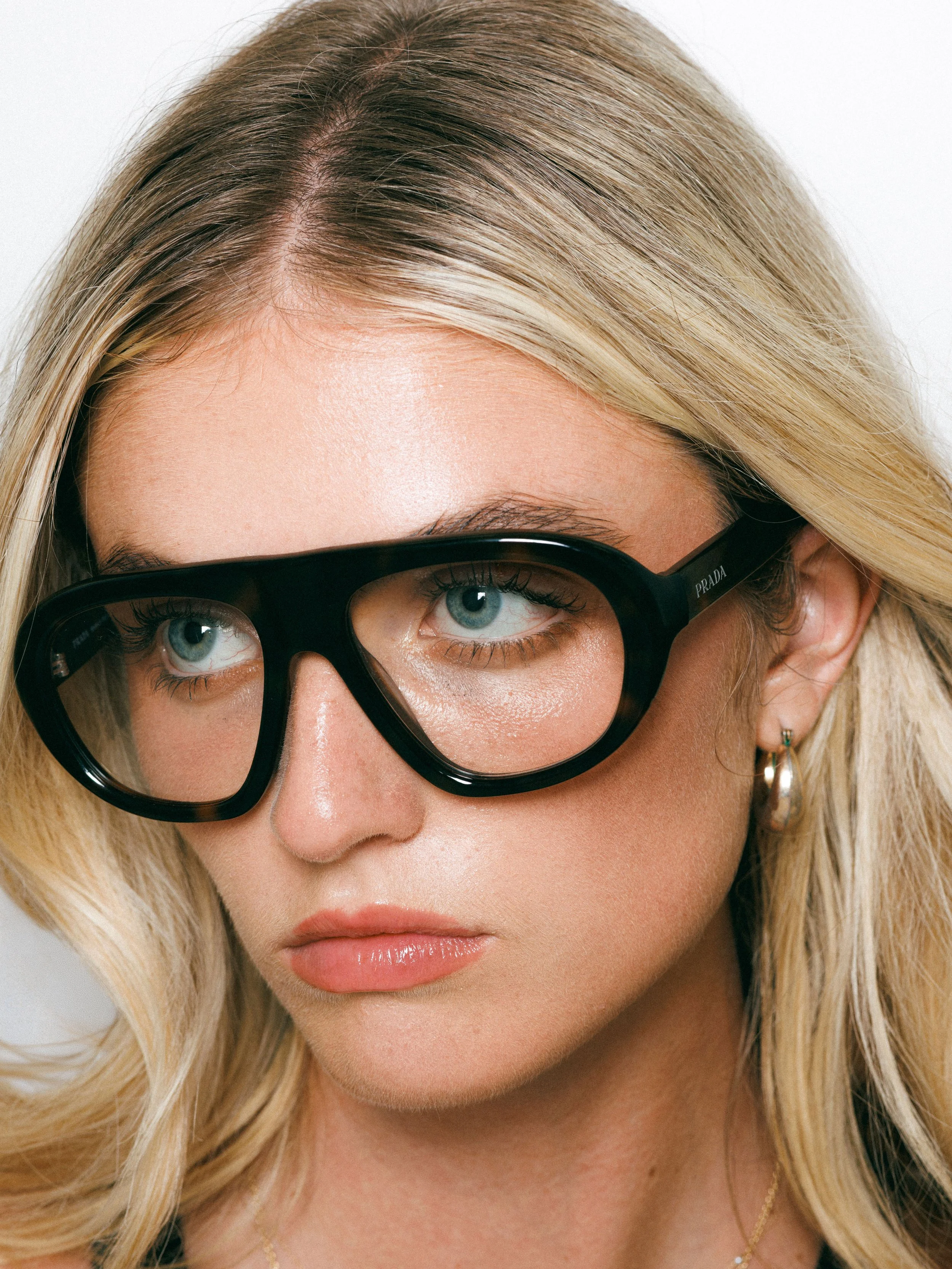 Close-up portrait of a blonde fashion model wearing oversized black glasses, gold hoop earrings, and a black sleeveless top against a white background.
