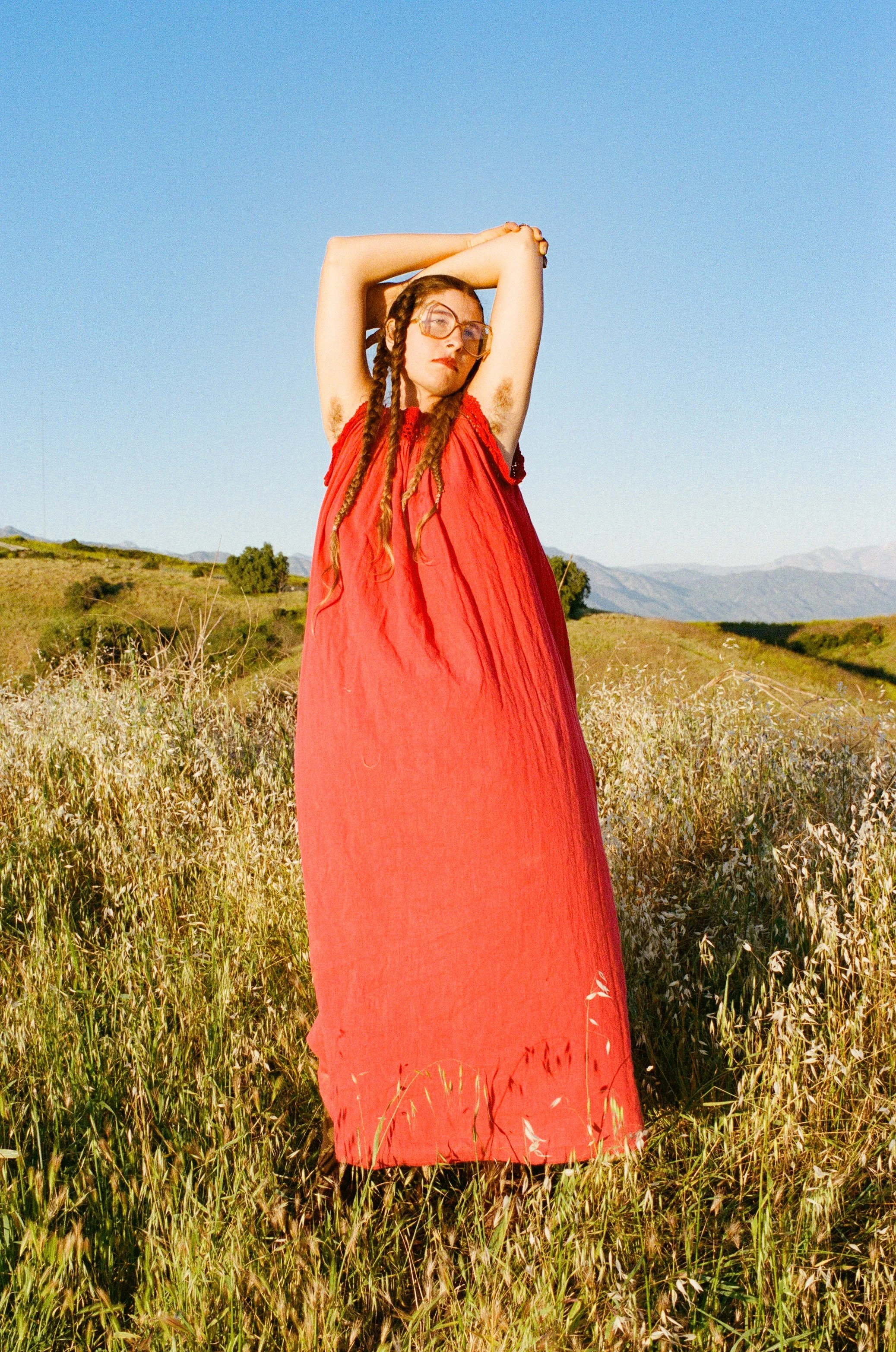 Woman with braided hair in a red dress standing in a field of tall grass under a clear blue sky with mountains in the distance.