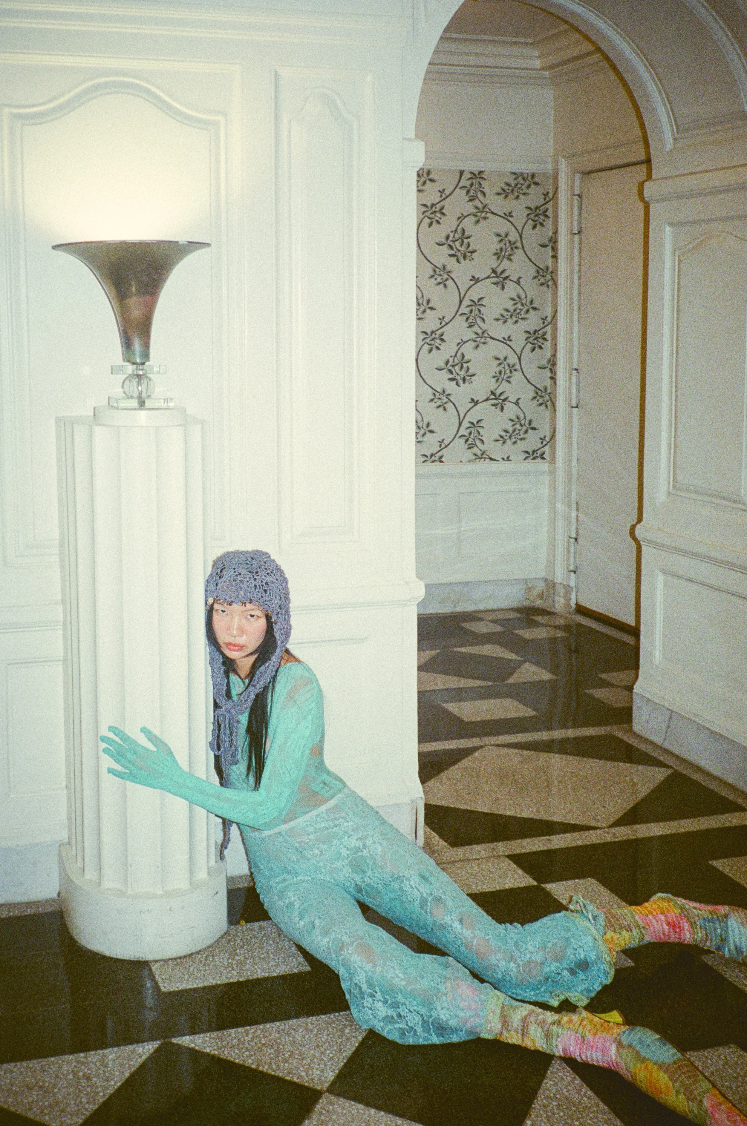 Fashion model wearing a colorful dress, lace stockings, and a knit hat, reclining on a black-and-white checkered floor against a white column with an artistically extended arm reaching upward.