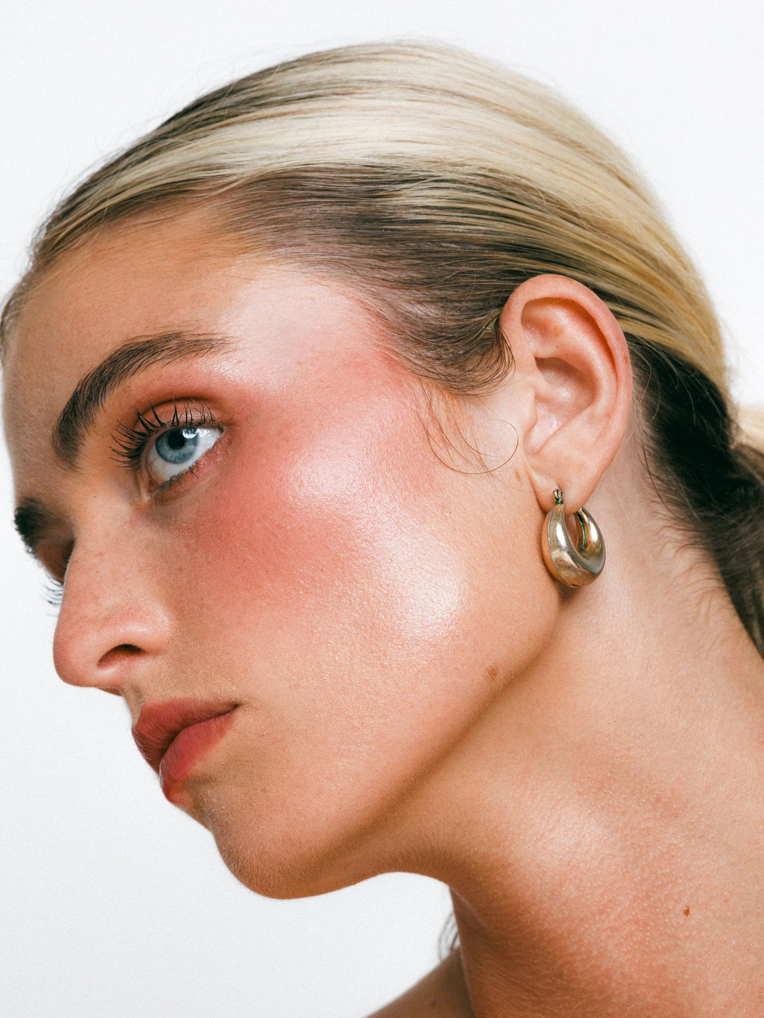 Close-up beauty portrait of a blonde model with glowing makeup, gold hoop earrings, and a sleek ponytail against a white background.