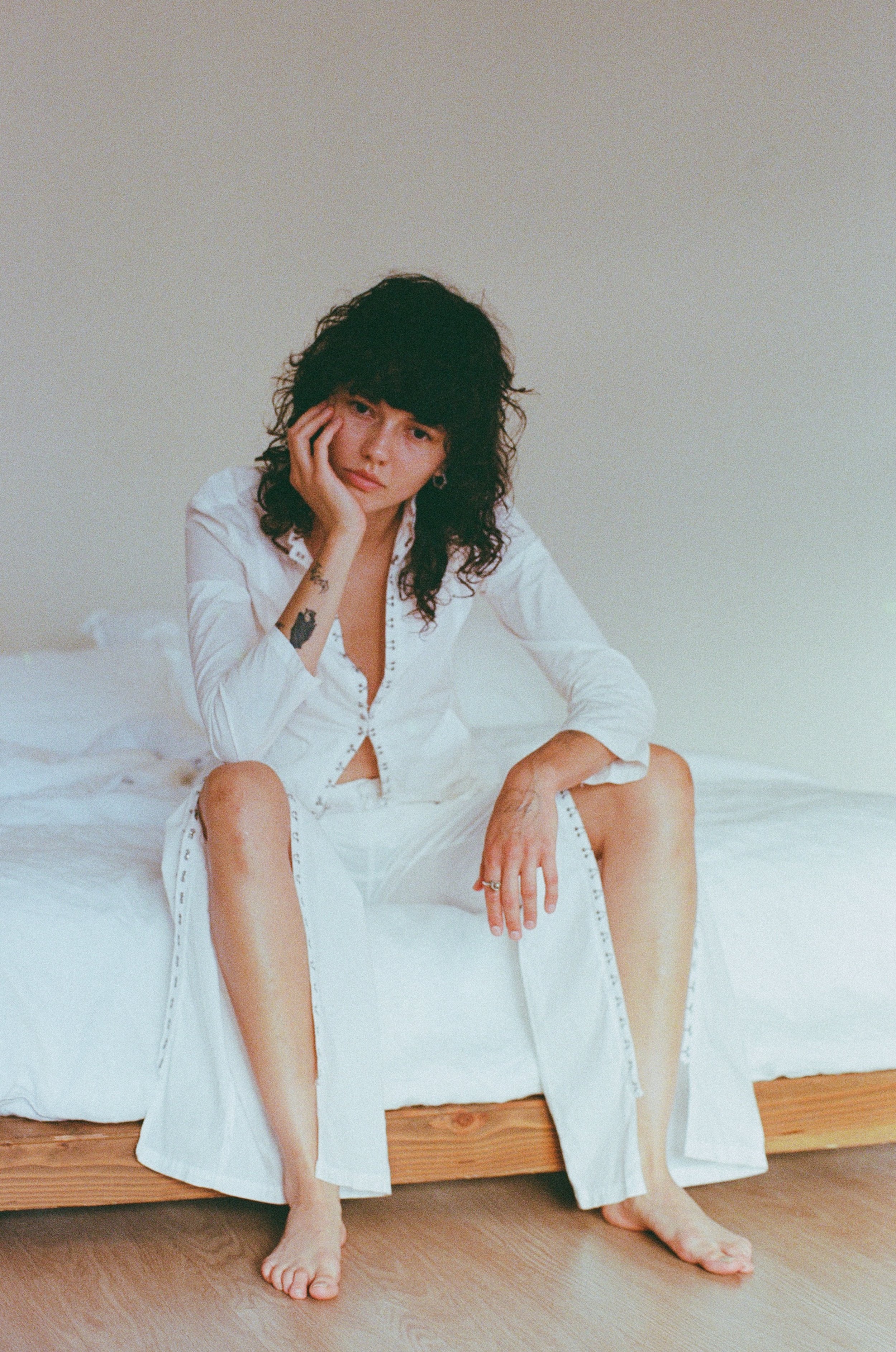 A woman with curly dark hair sitting on a bed, wearing a long white shirt, with her hand on her face and legs apart, with tattoos visible on her arm.