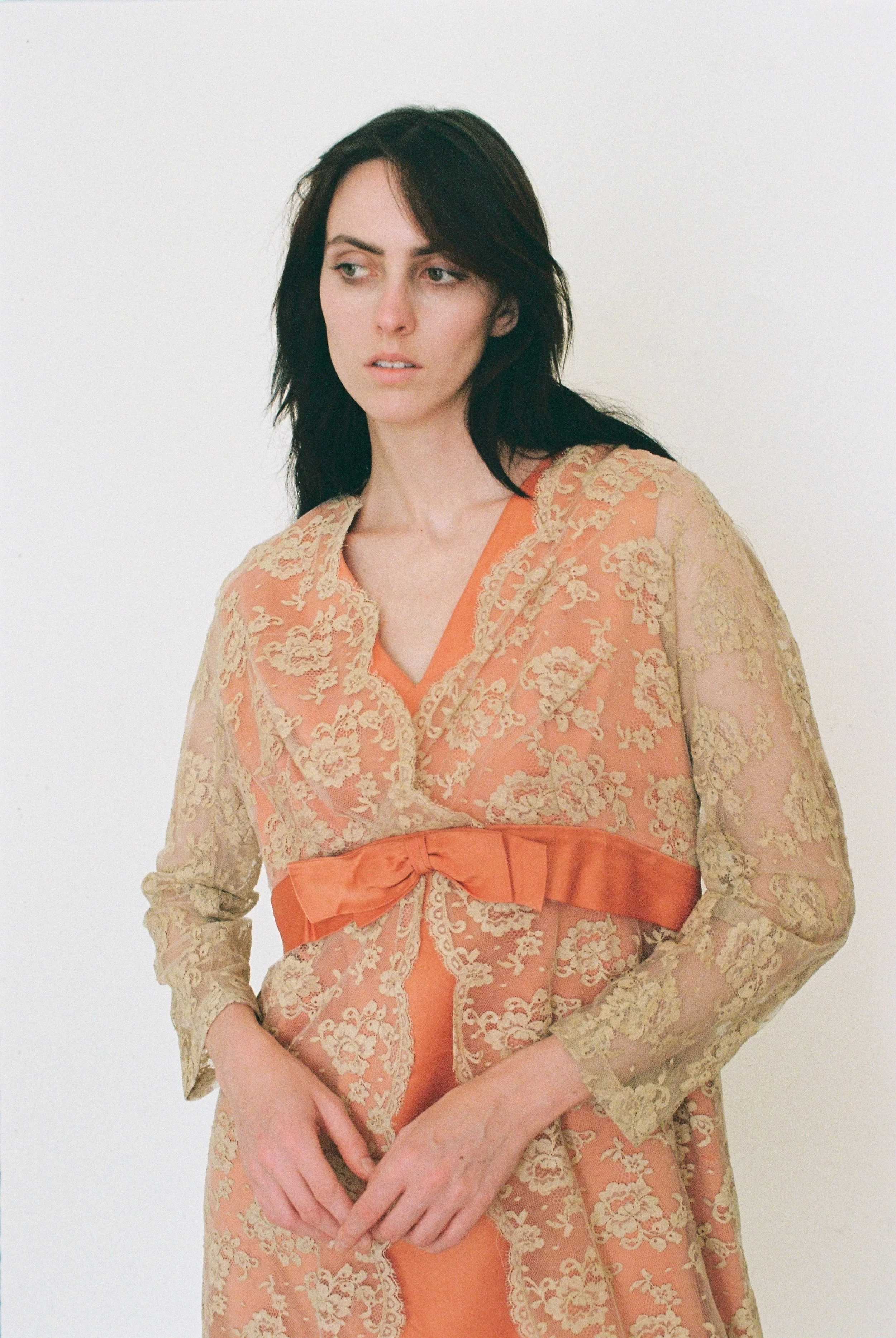 Fashion model with dark hair wearing an orange dress with a beige lace overlay and a large bow at the waist, standing against a white background.