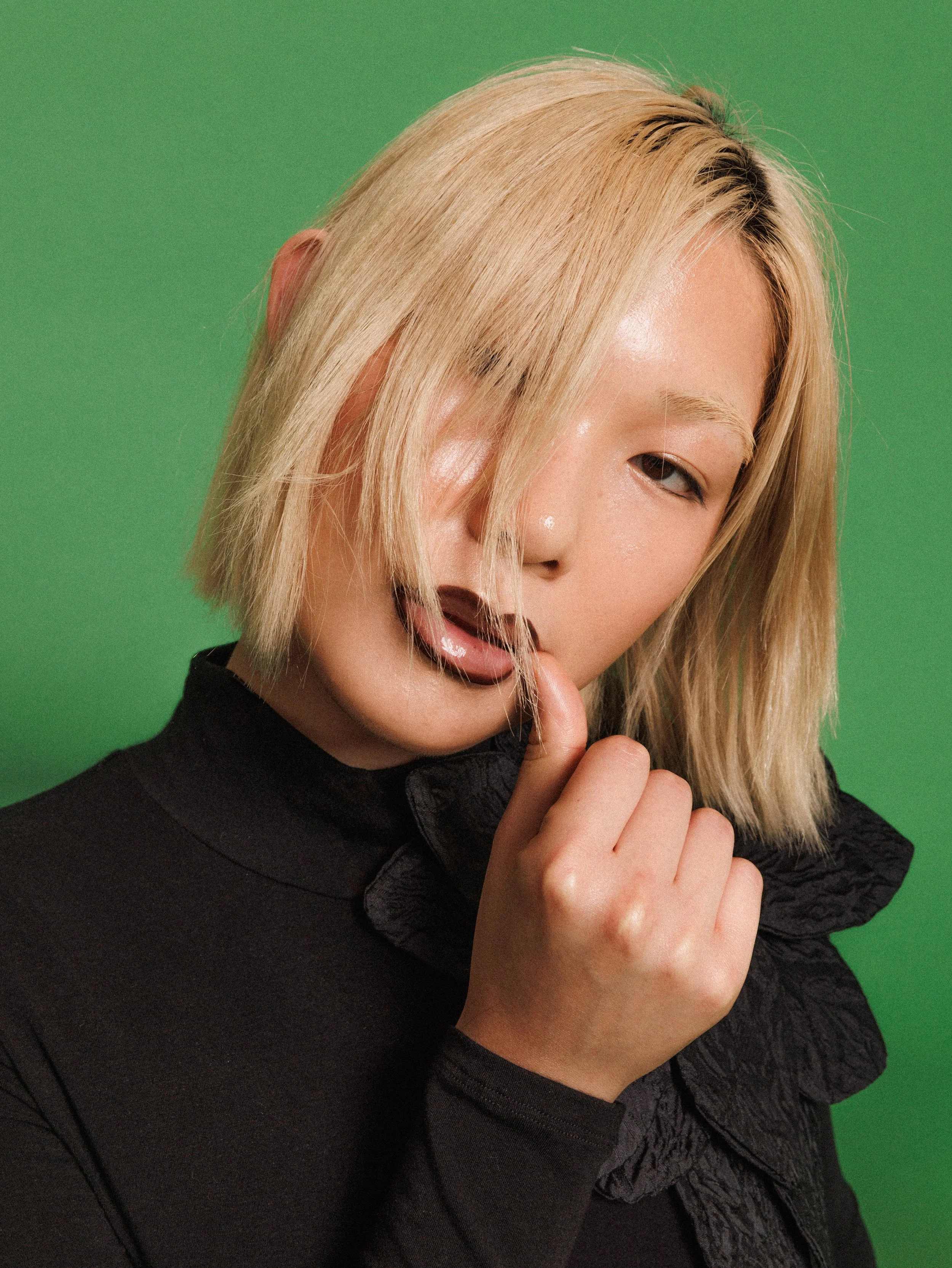 Editorial fashion portrait of a model wearing a black long-sleeve top with an oversized flower, photographed against a vibrant green studio background.