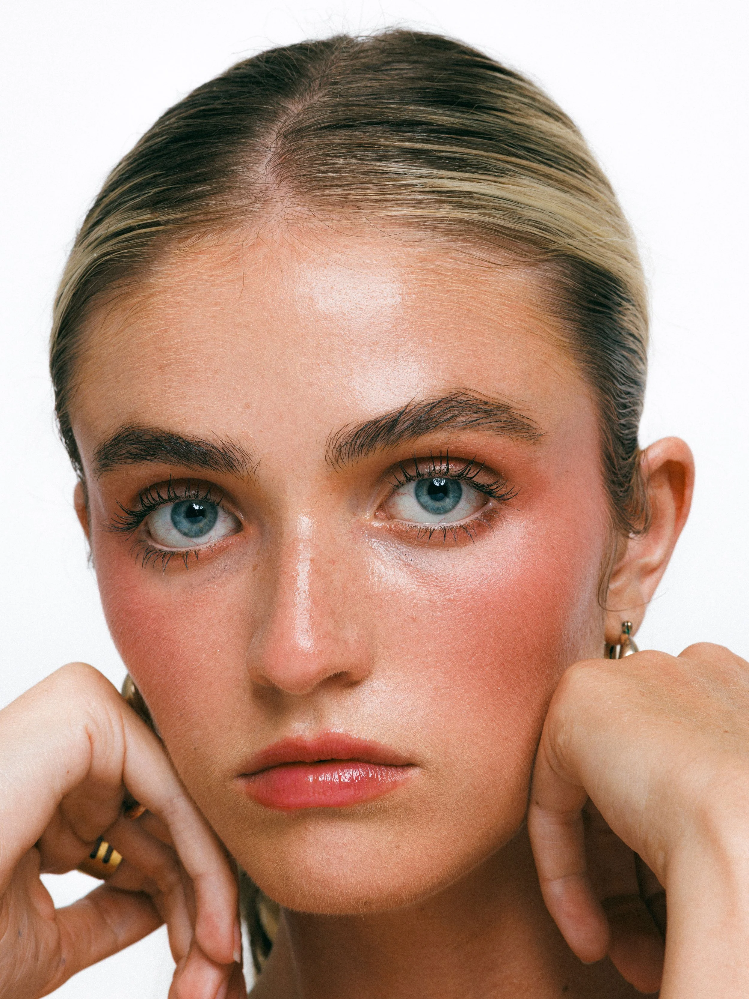 Close-up beauty portrait of a blonde model with glowing makeup, gold hoop earrings, and a sleek ponytail against a white background.