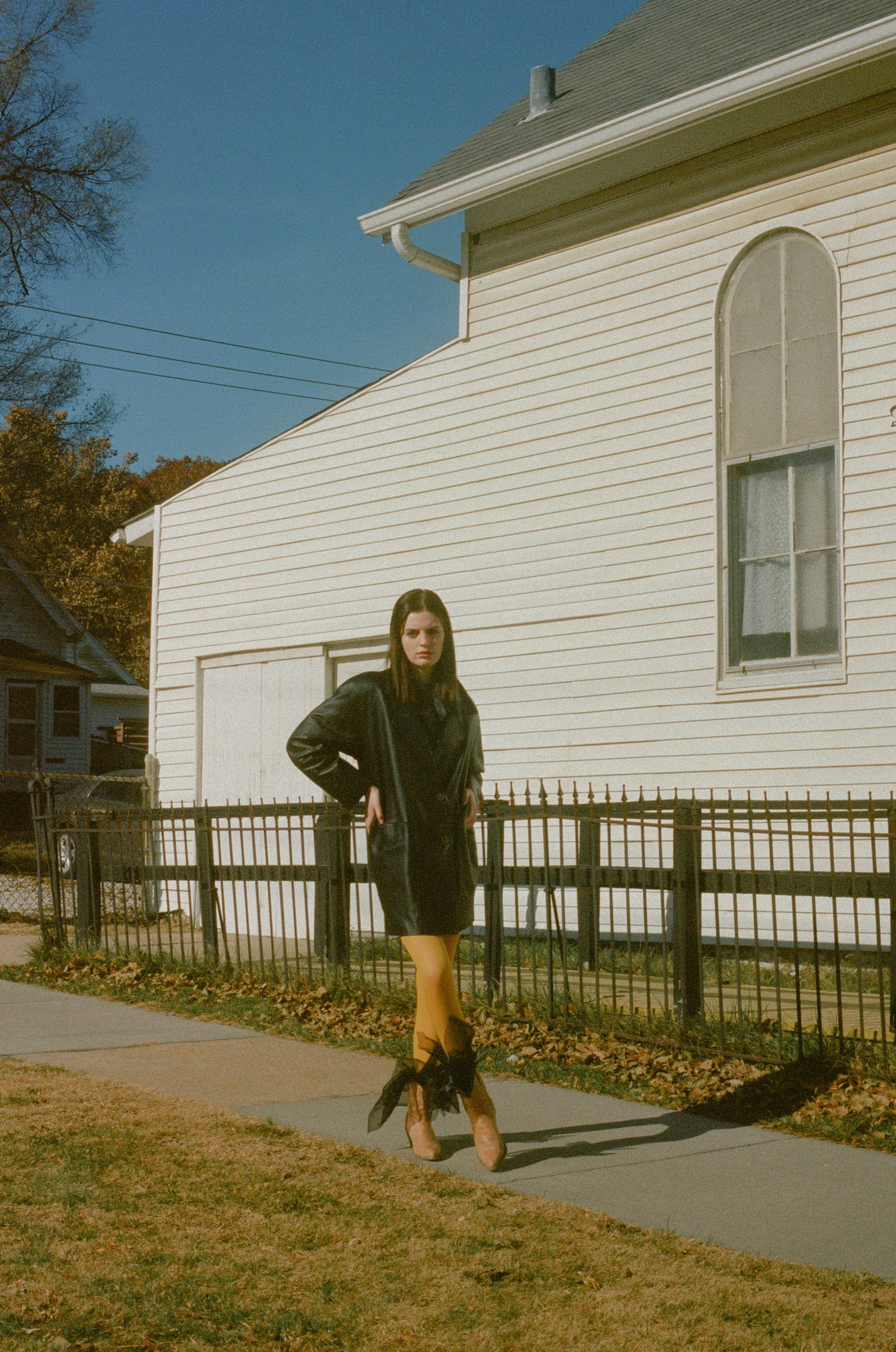 Fashion model wearing a black leather jacket, yellow leggings, and high heels, standing outside near a white house with arched windows.