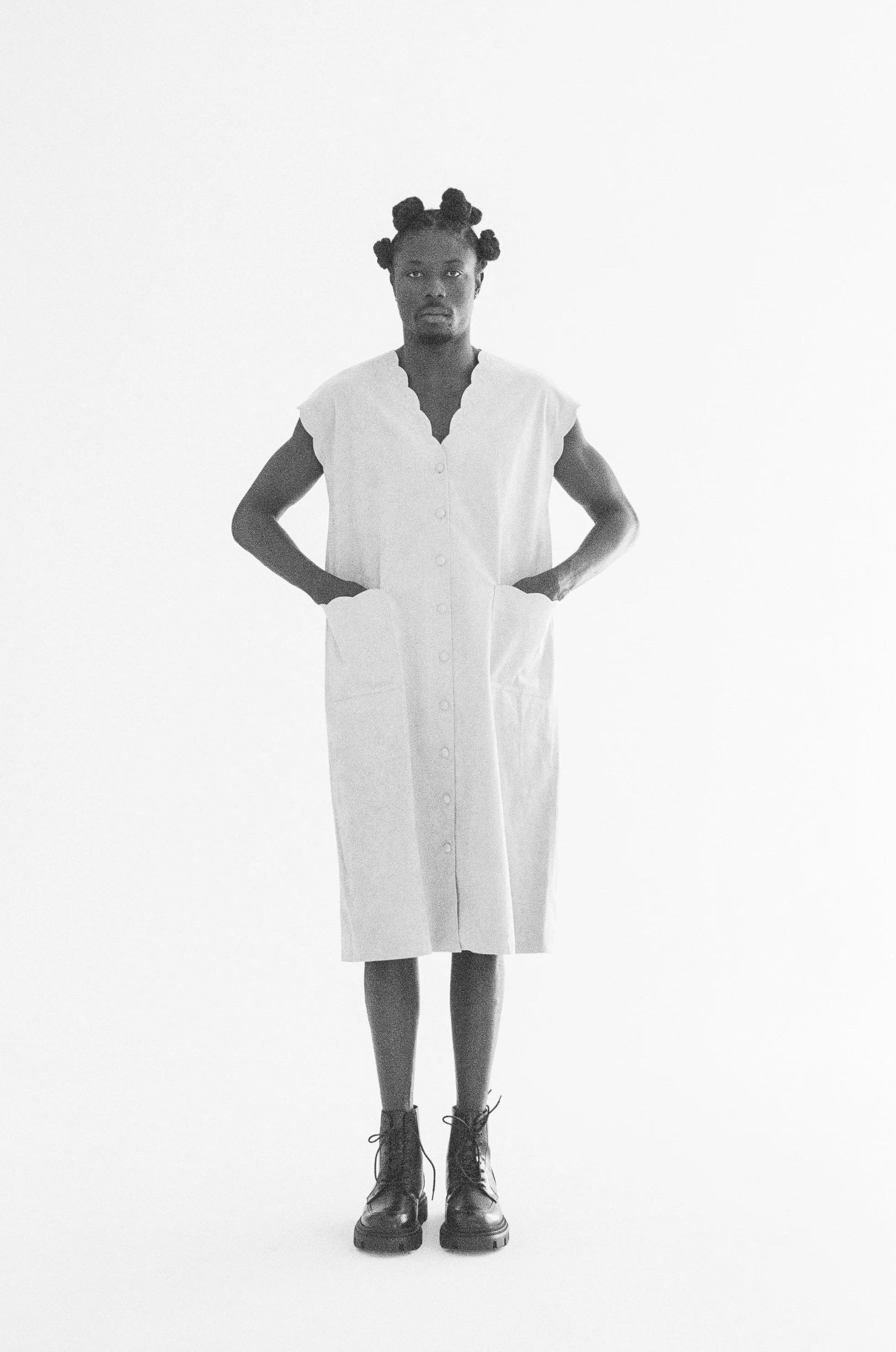 Fashion model standing with his hands in his pockets, wearing a sleeveless button-front dress and black boots against a white background.