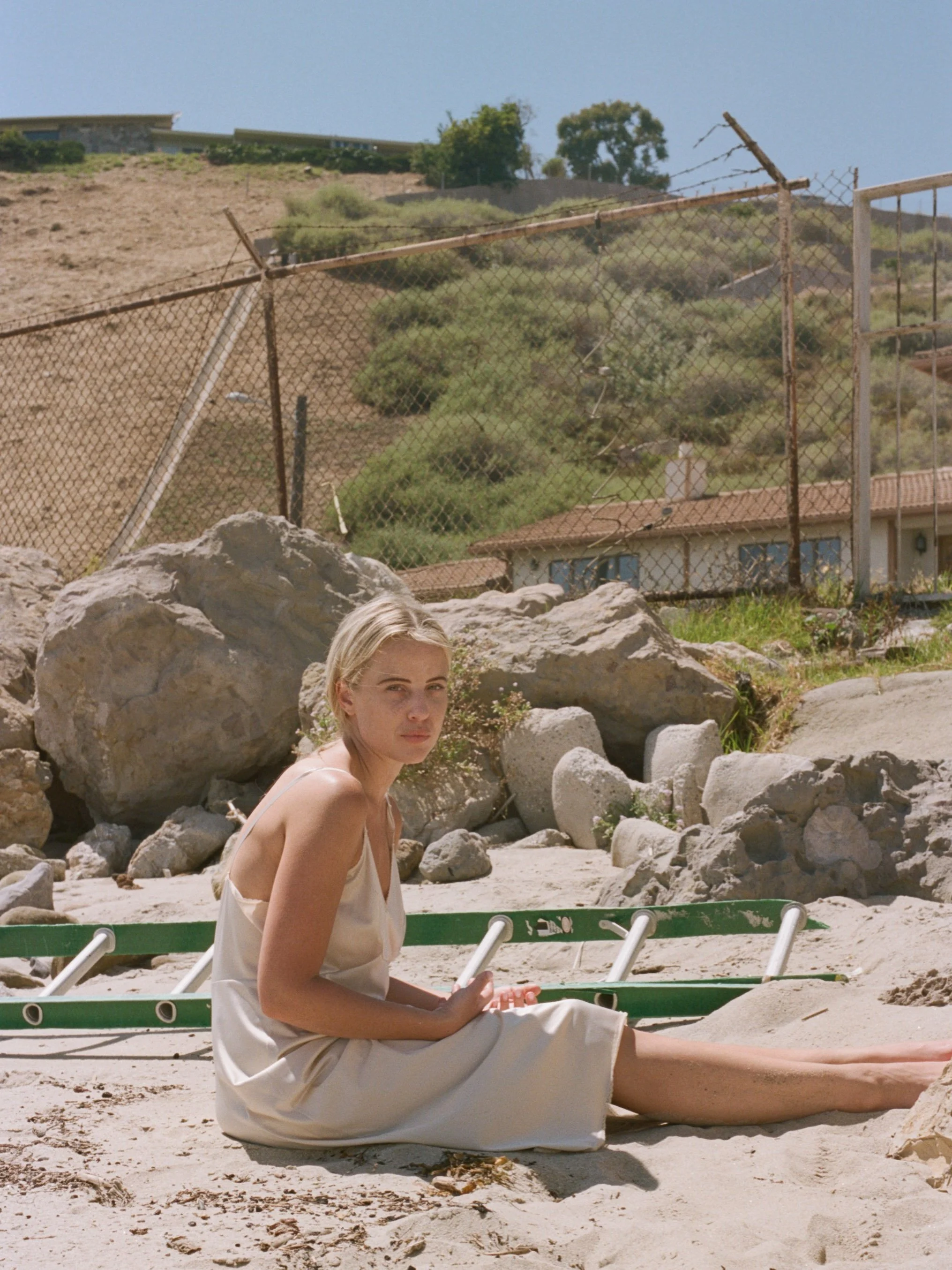 A young woman in a white dress sitting on sandy ground at the beach, surrounded by rocks, holding a phone, with a green life-saving ring nearby, and a rock formation behind her.