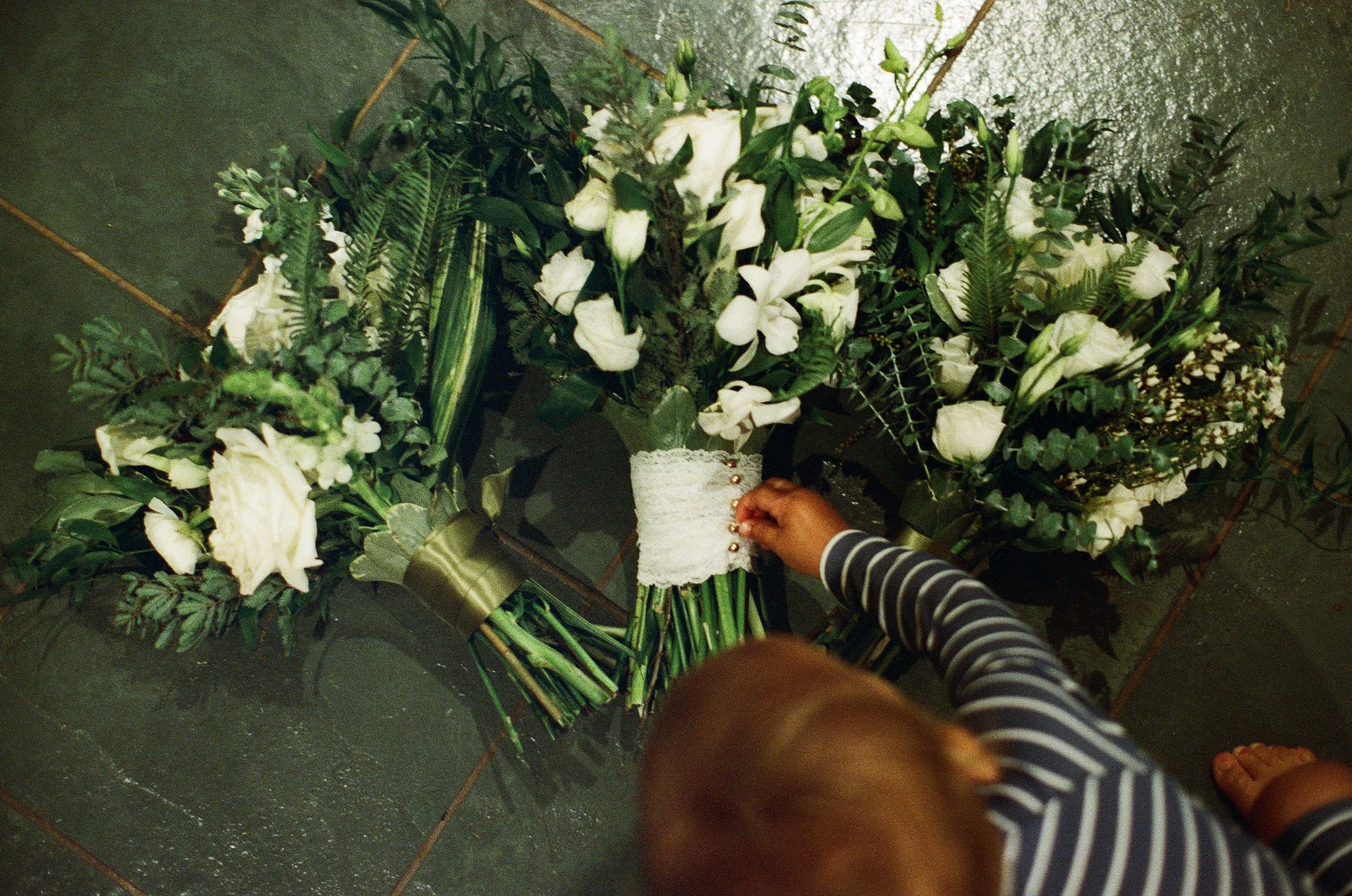 A toddler grasping for a white lace bouquet, surrounded by large white flowers and greenery, on a dark surface.
