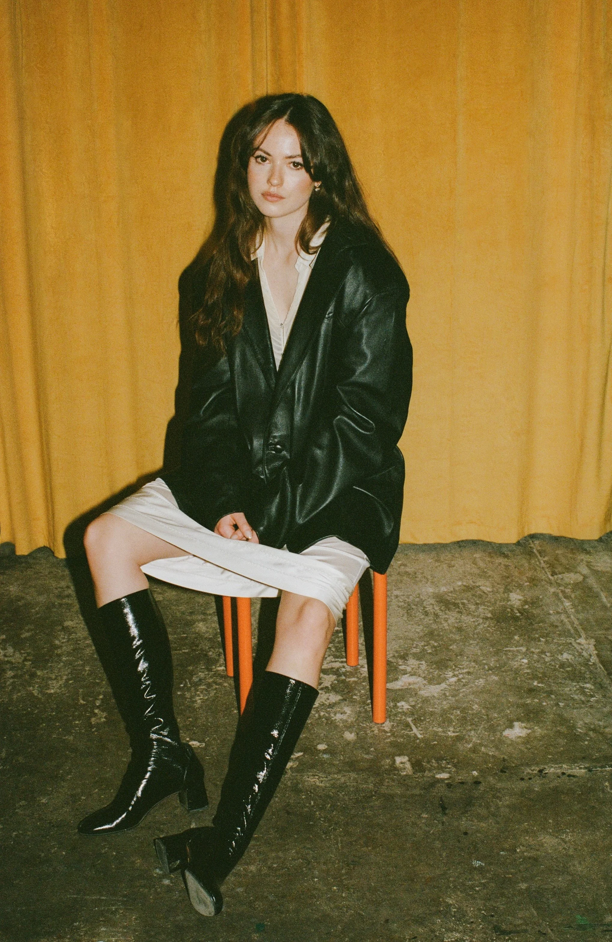 Woman in a black leather jacket and boots seated on a chair against a warm gold backdrop.