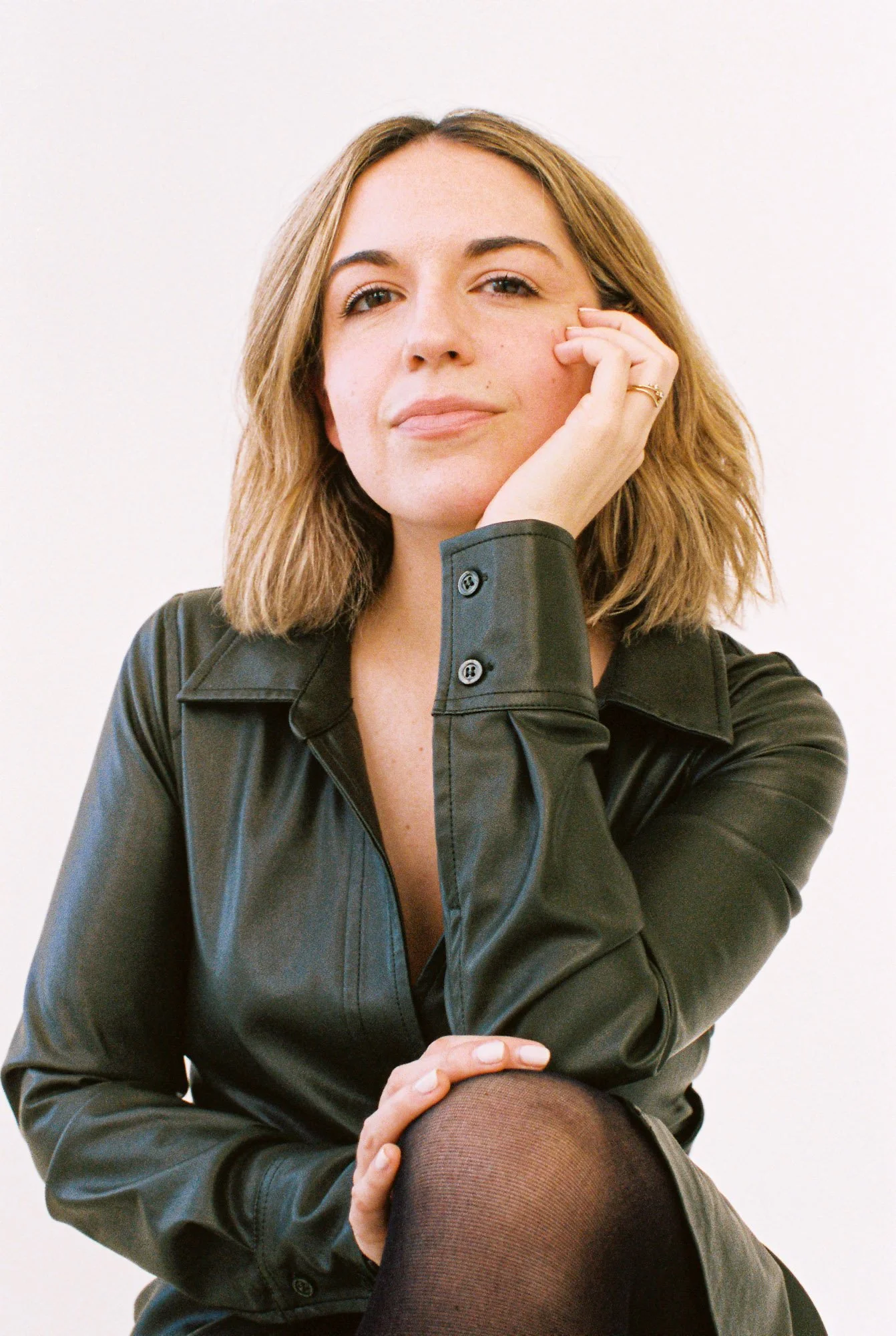 Professional portrait of a woman with shoulder-length blonde hair wearing a black leather top, seated with her hand resting against her face against a clean light background.