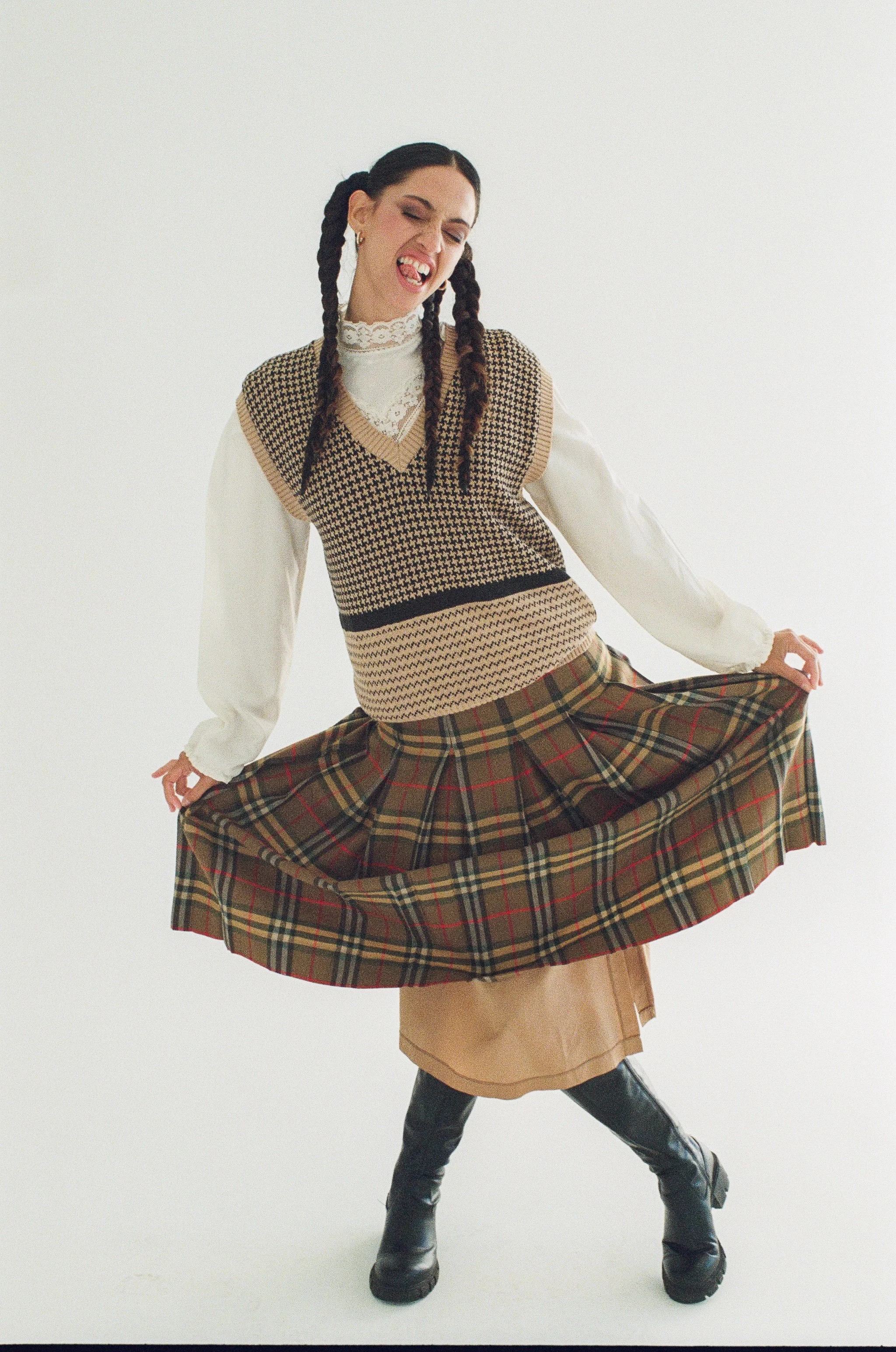 Fashion model with braided hair wearing a knitted vest over a lace-collared blouse, a plaid skirt, and black boots against a white background.