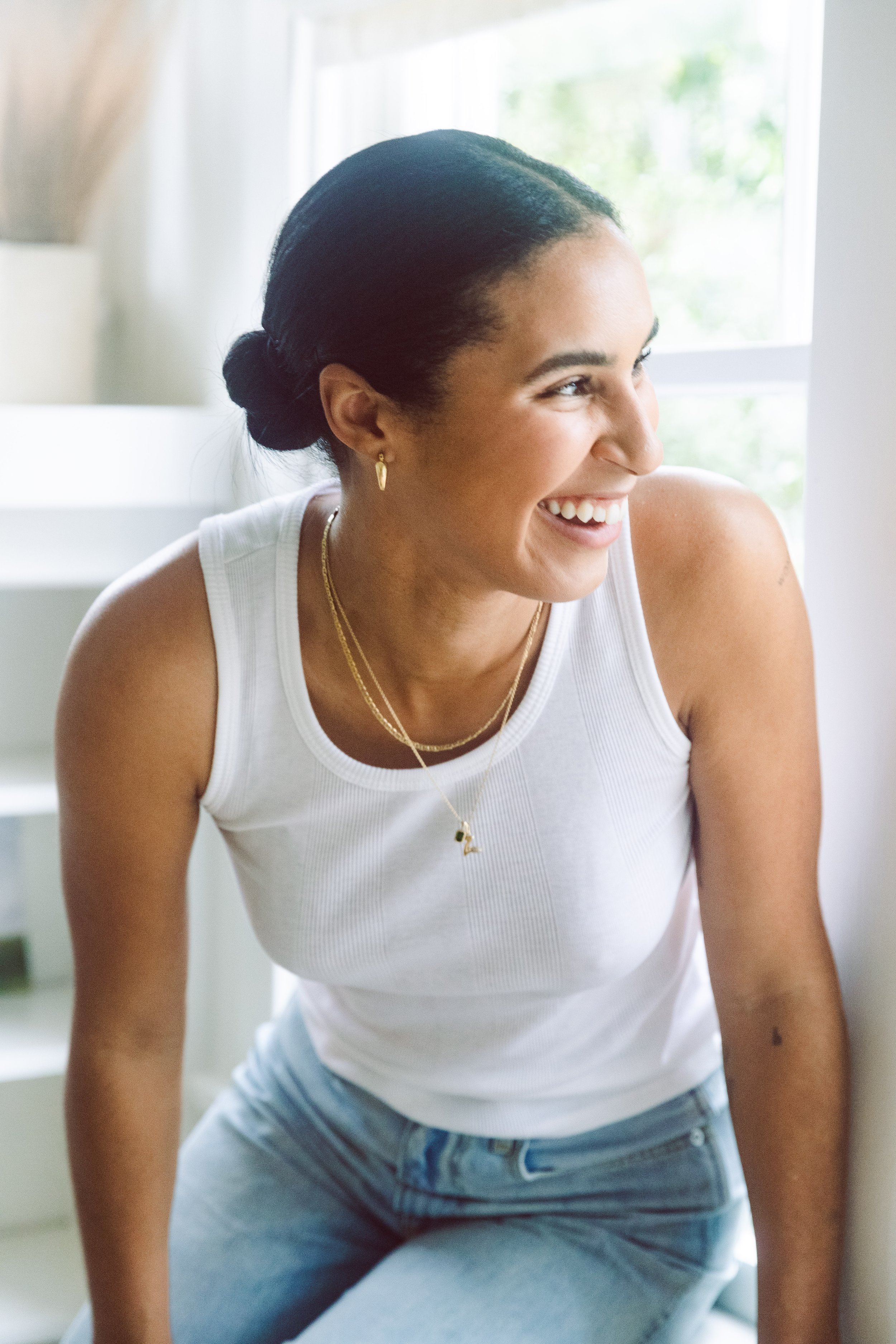Smiling model in a white tank top and jeans seated by a bright window.