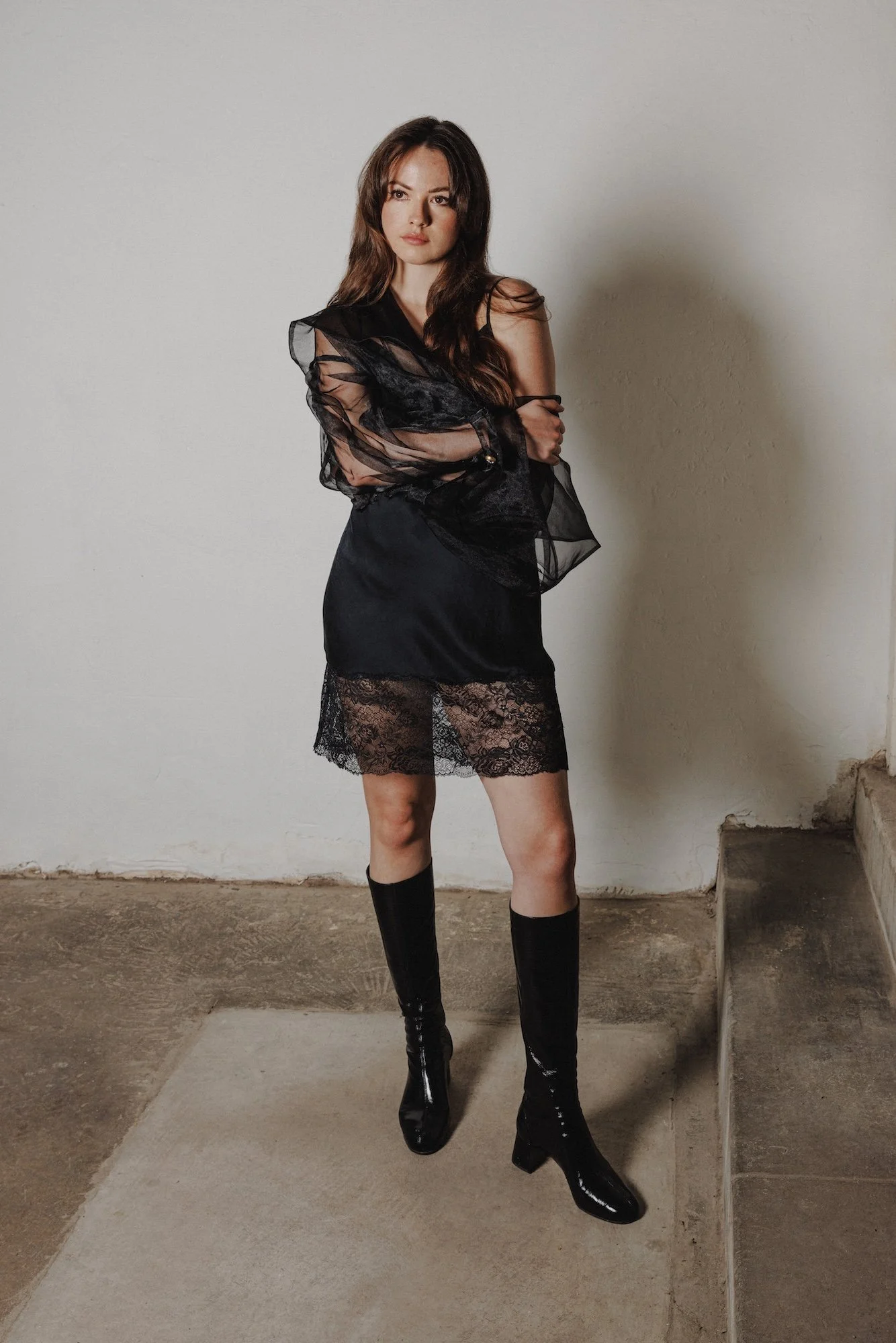 A woman with long brown hair, wearing a black lace skirt, black high-heeled boots, and a sheer black top with ruffled sleeves, standing against a plain beige wall with a concrete floor.