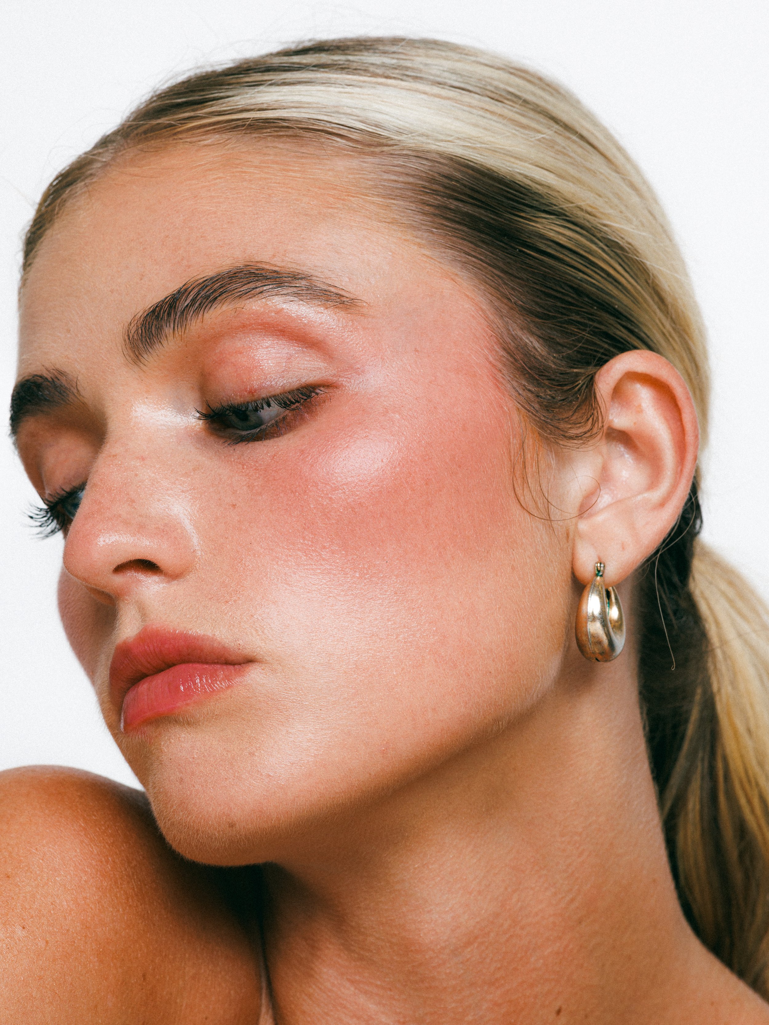Close-up beauty portrait of a blonde model with glowing makeup, gold hoop earrings, and a sleek ponytail against a white background.