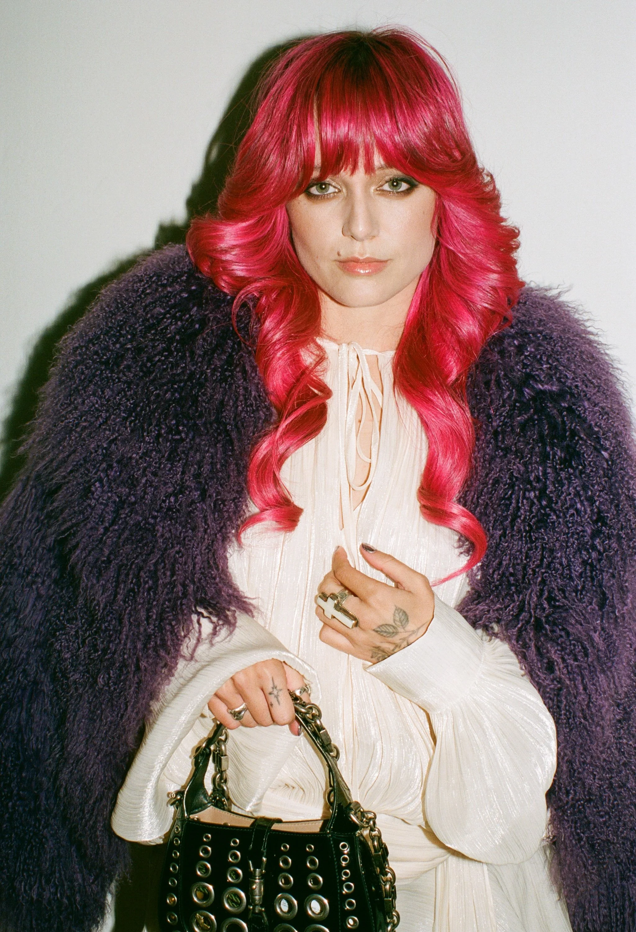 A woman with bright pink hair styled in loose waves, wearing a purple fuzzy coat over a cream-colored, pleated dress, holding a black handbag with silver grommets and chain strap, standing against a plain white background.