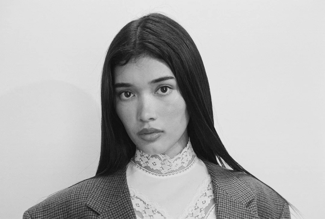Black and white portrait of a young woman with straight dark hair, wearing a lace high-neck top and a plaid blazer, against a plain light background.