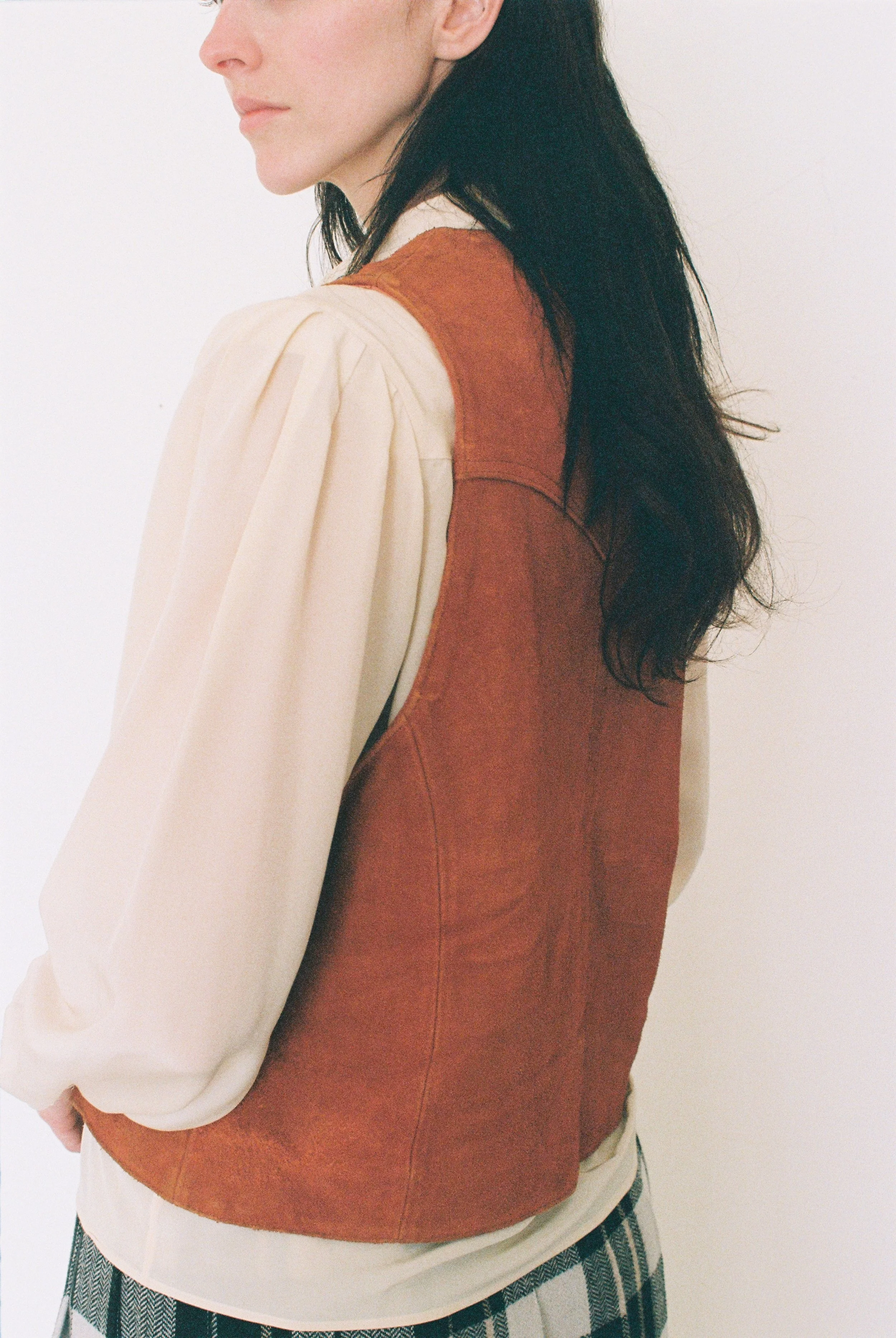 Partial side view of a fashion model wearing a beige blouse and a brown leather vest against a white background.