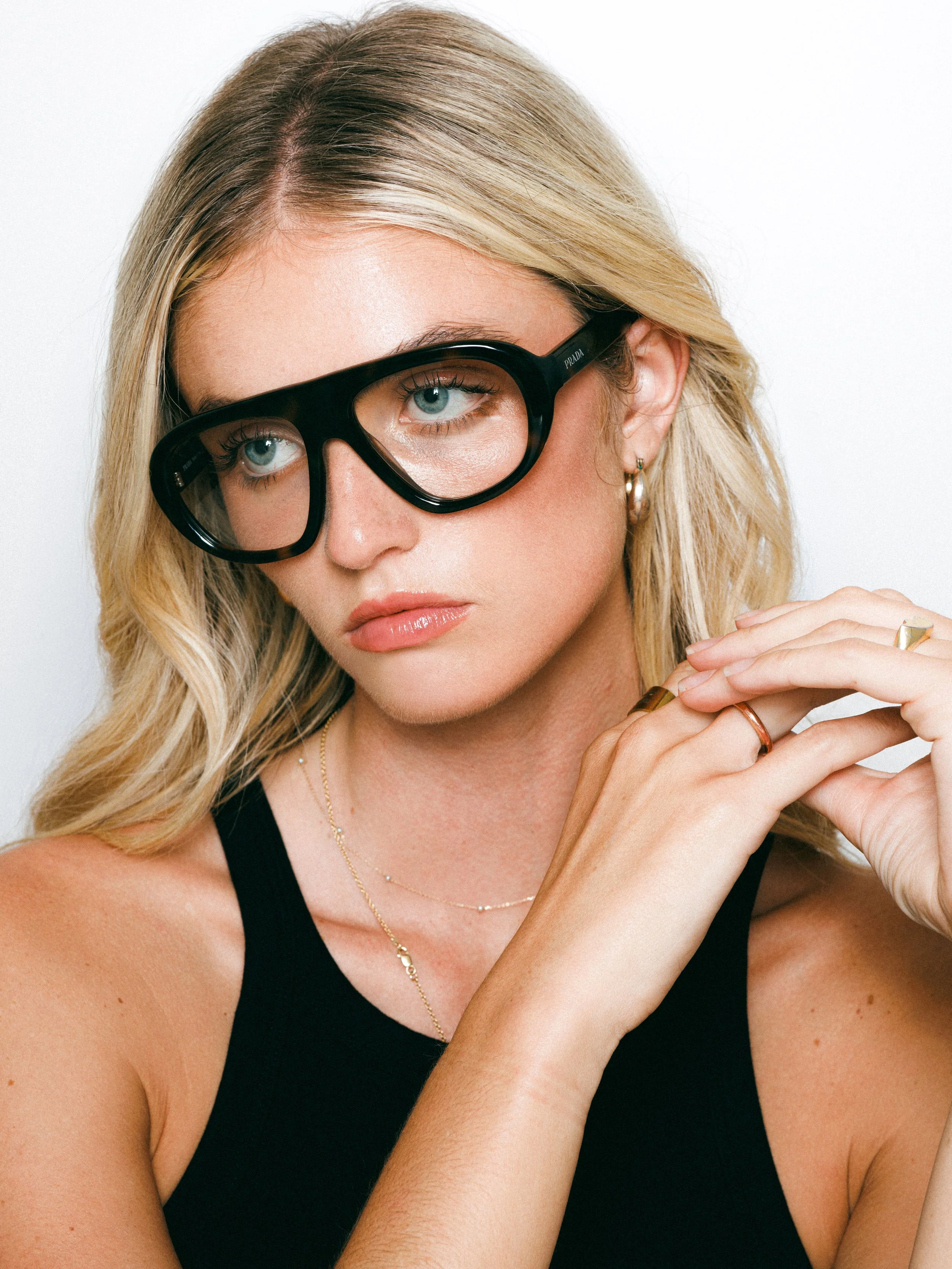 Close-up portrait of a blonde fashion model wearing oversized black glasses, gold hoop earrings, and a black sleeveless top against a white background.