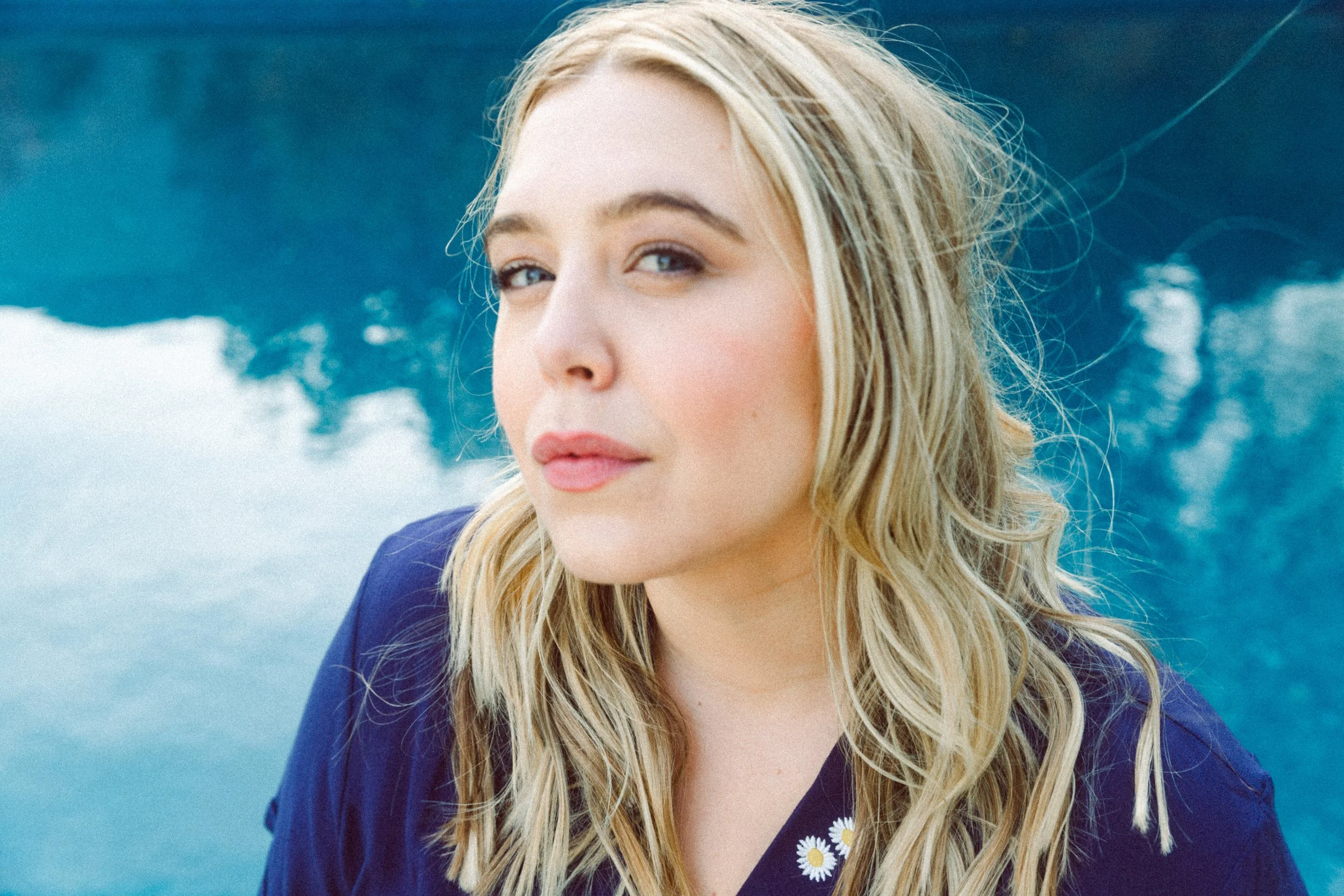 A woman with blonde hair and blue eyes looking at the camera. She is wearing a blue coat, and there are daisy flowers sewn on the front of her shirt. The background includes trees and a partly cloudy sky.