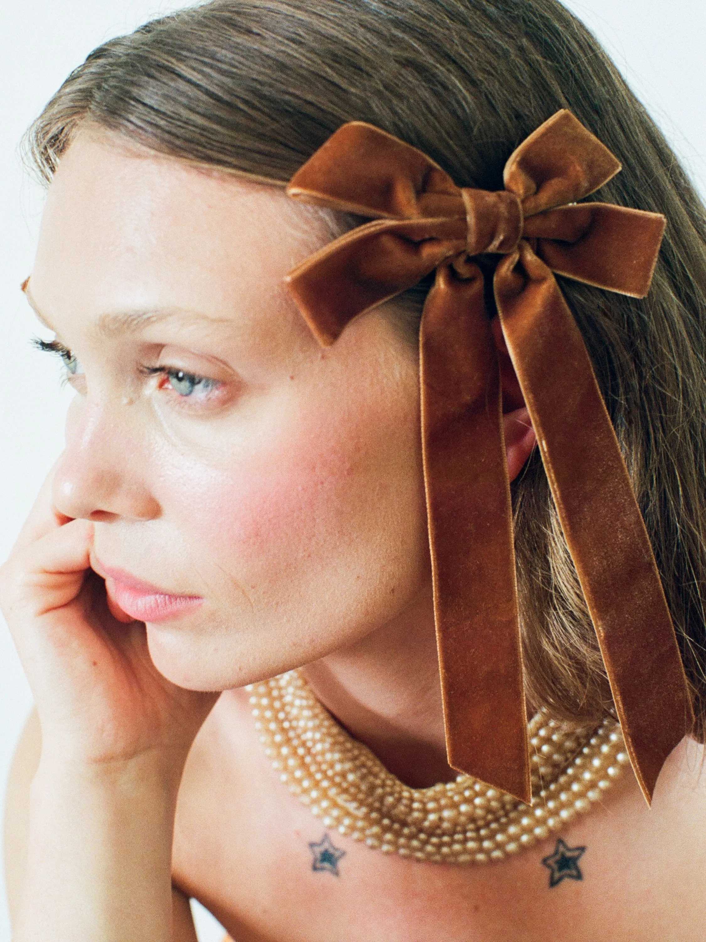 Fashion model with styled brown hair wearing a large brown velvet bow, a pearl necklace, and star tattoos on her chest, resting her chin on her hand.
