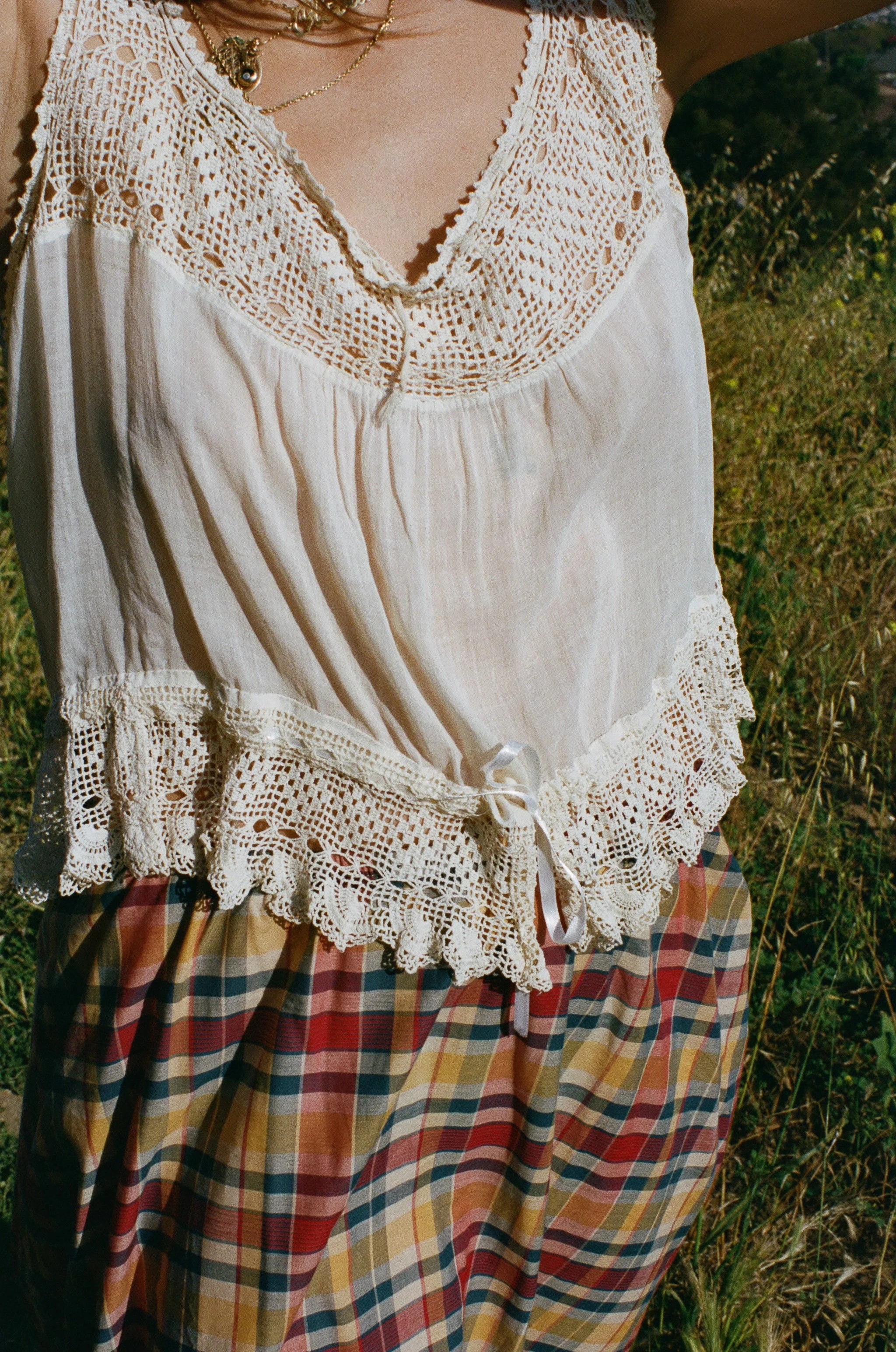 Close-up of a fashion model wearing a white lace blouse and a red, yellow, and blue plaid skirt, photographed outdoors with grass in the background.
