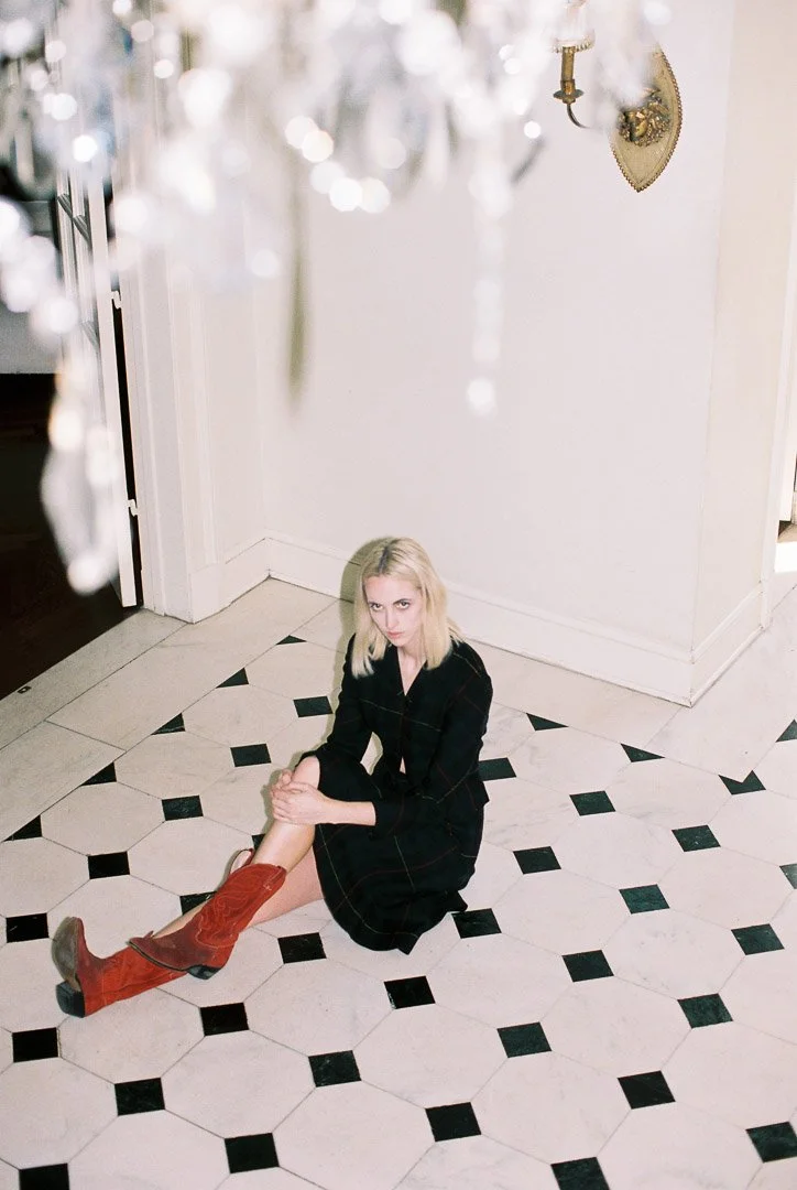 Editorial fashion photograph of a model wearing a dark outfit and red knee-high boots, seated on a black-and-white tiled floor beneath a crystal chandelier.
