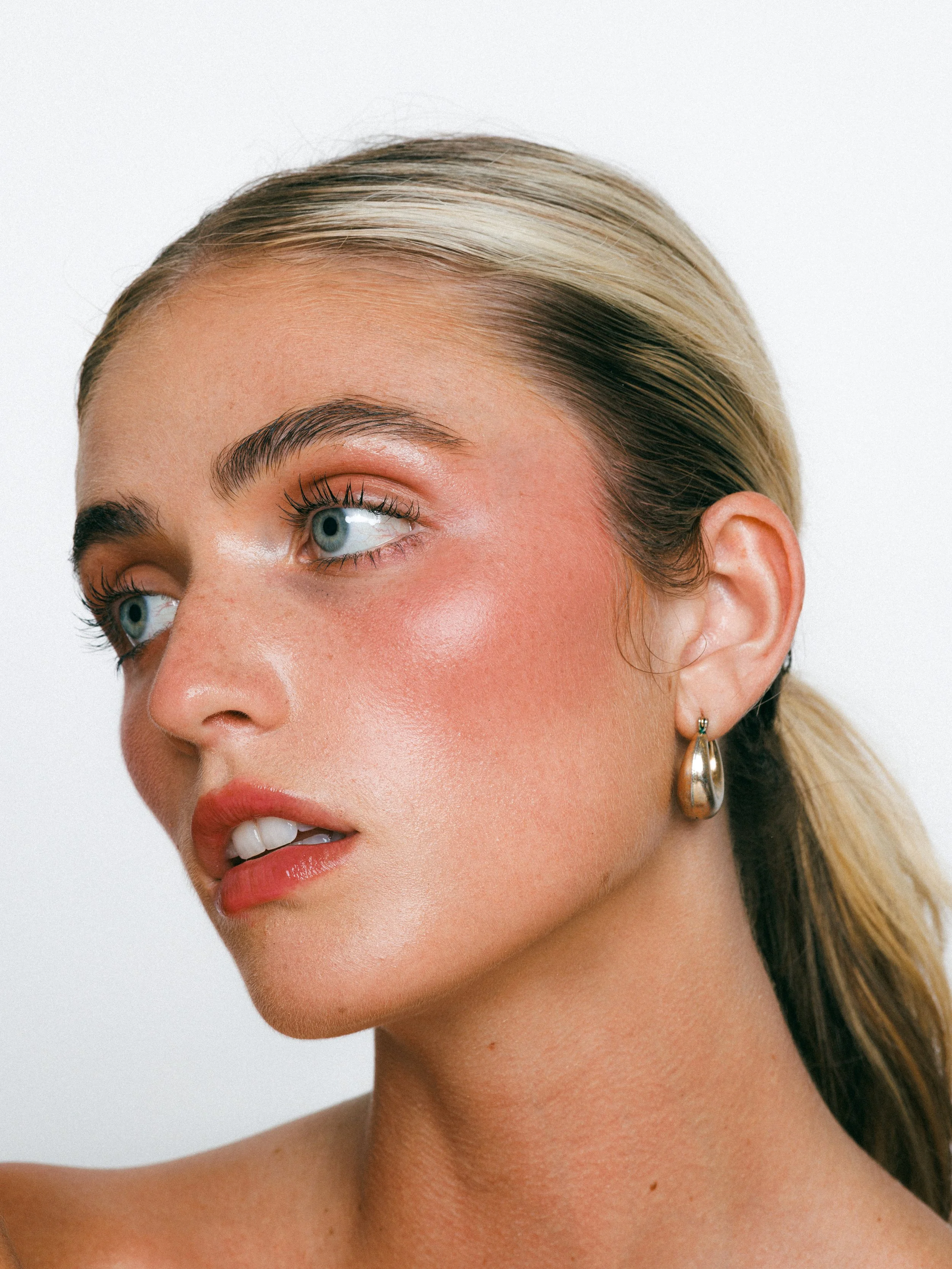 Close-up beauty portrait of a blonde model with glowing makeup, gold hoop earrings, and a sleek ponytail against a white background.