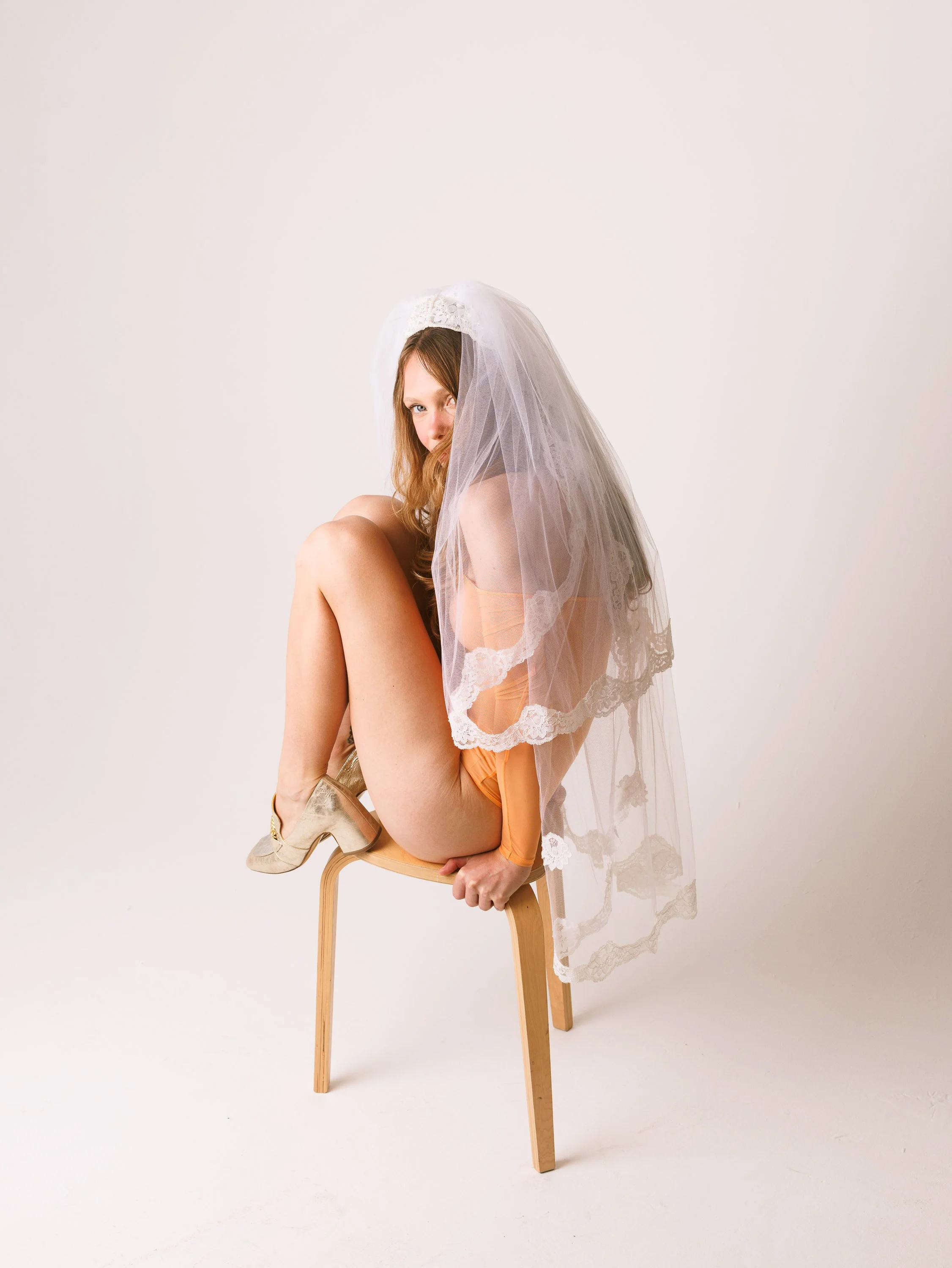 Fashion model with long hair sitting on a wooden chair wearing a bridal veil and an orange mesh bodysuit, holding the chair legs with one knee raised.