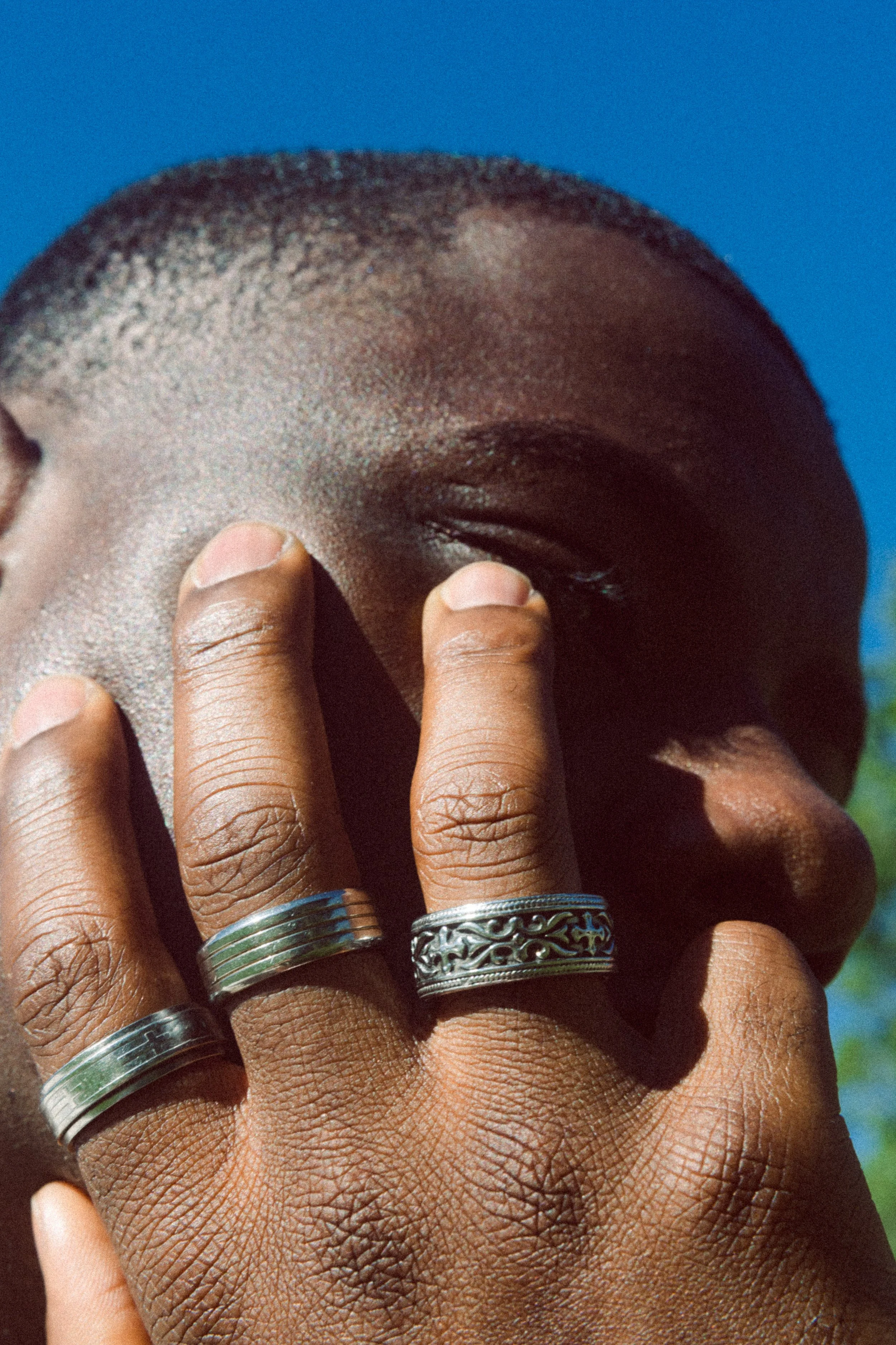 Close-up of a fashion model’s hand wearing four rings, resting against the face against a blue background.