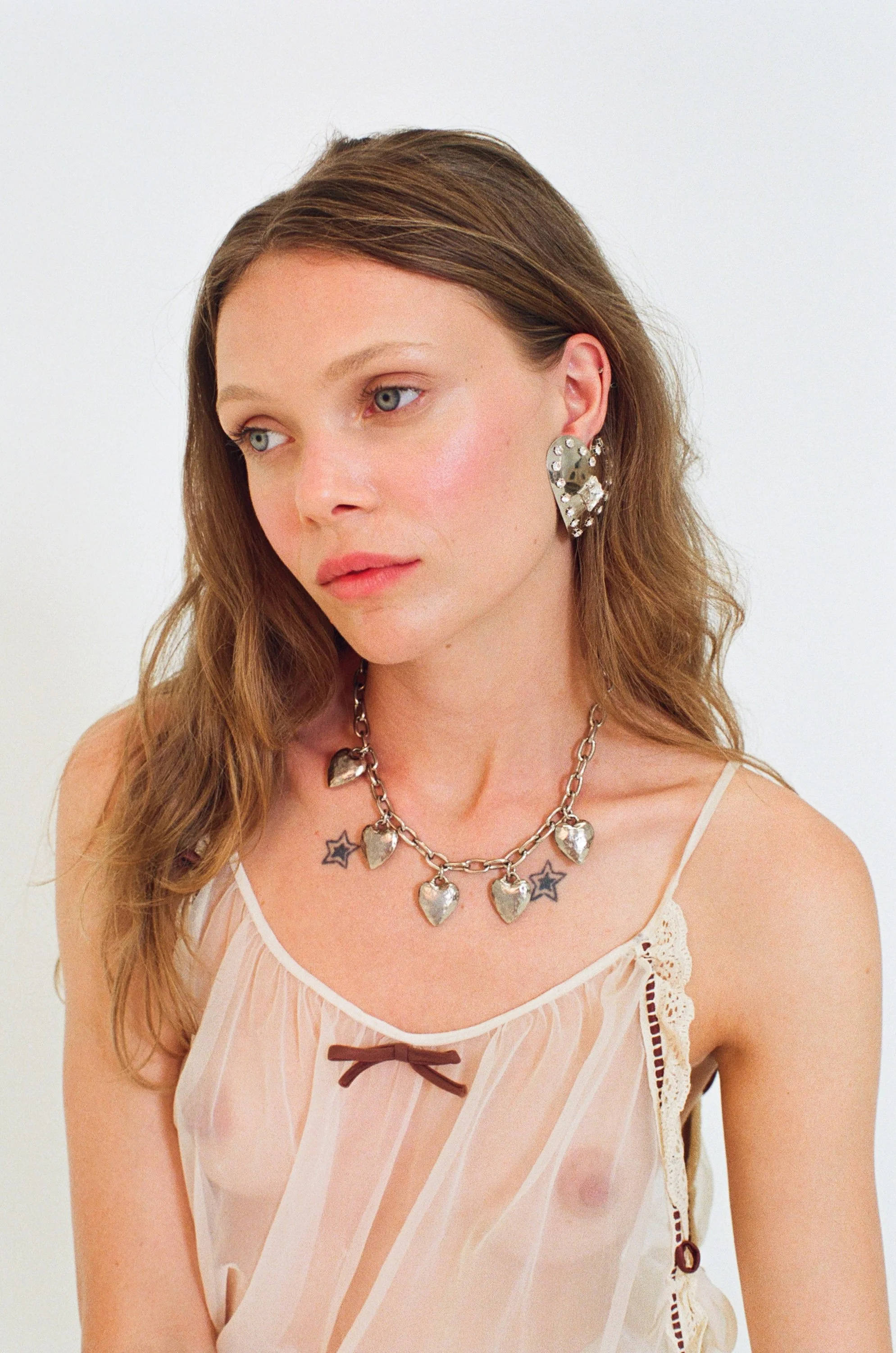 Fashion model with long wavy hair wearing a cream-colored lace-trimmed top, large silver heart-shaped earrings, a heart charm necklace, and visible collarbone tattoos.