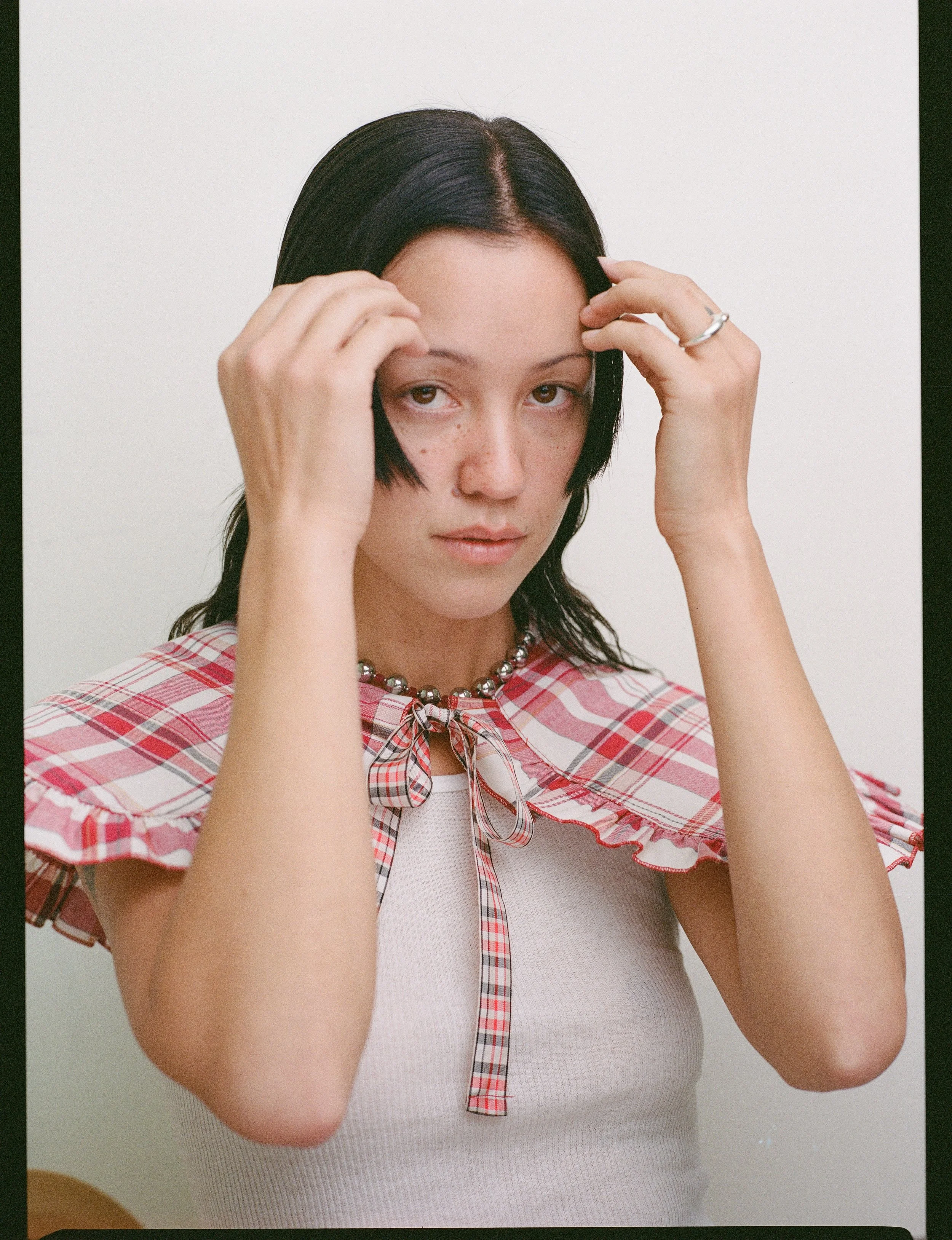 A woman with black hair and freckles wears a white tank top and a red plaid blouse with ruffled sleeves and a bow detail, accessorized with a silver beaded necklace, and touches her forehead with her hands against a plain white background.