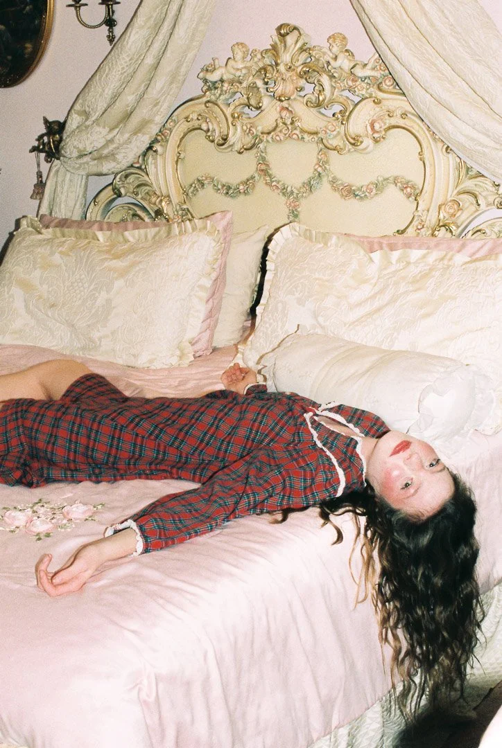 Fashion model with long dark curly hair wearing a red plaid pajama set, reclining on an ornate vintage bed with pink bedding and cream-colored pillows.