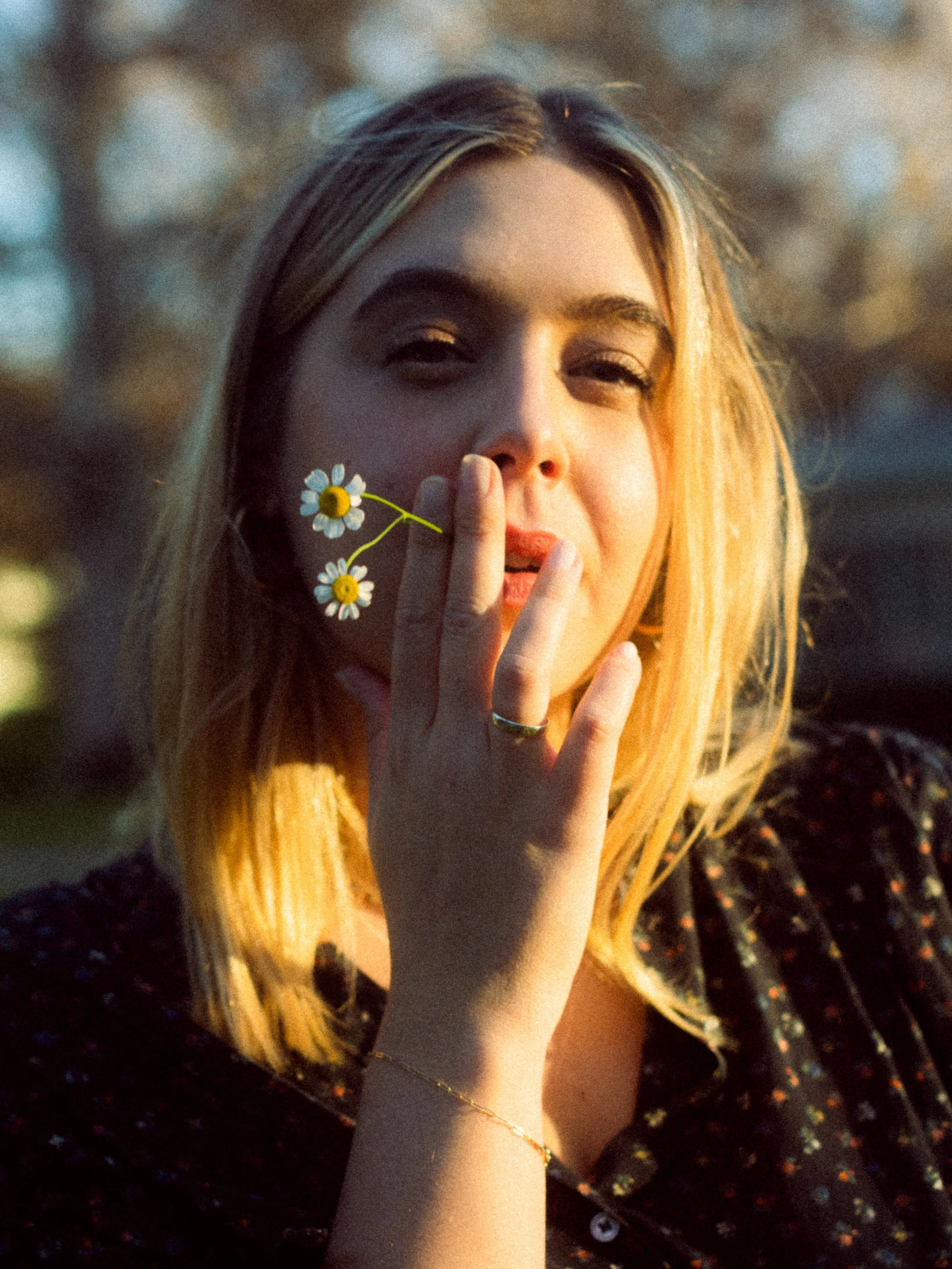 A young woman with long blonde hair, holding a small daisy flower near her face, with a blurred natural background, smiling gently.