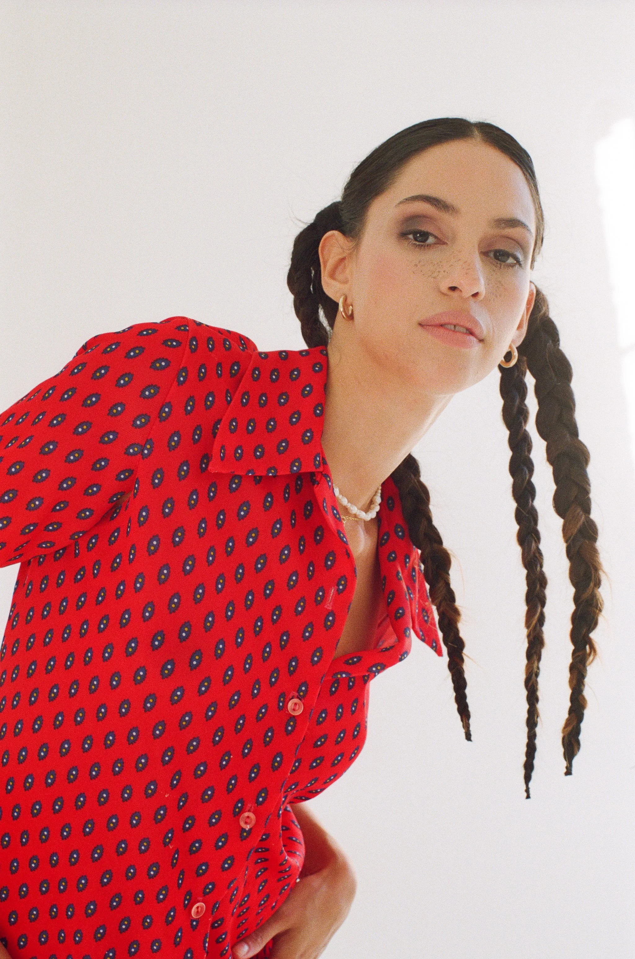 Model with long, dark braided hair wearing a red patterned shirt, gold hoop earrings, and a pearl necklace against a light background.
