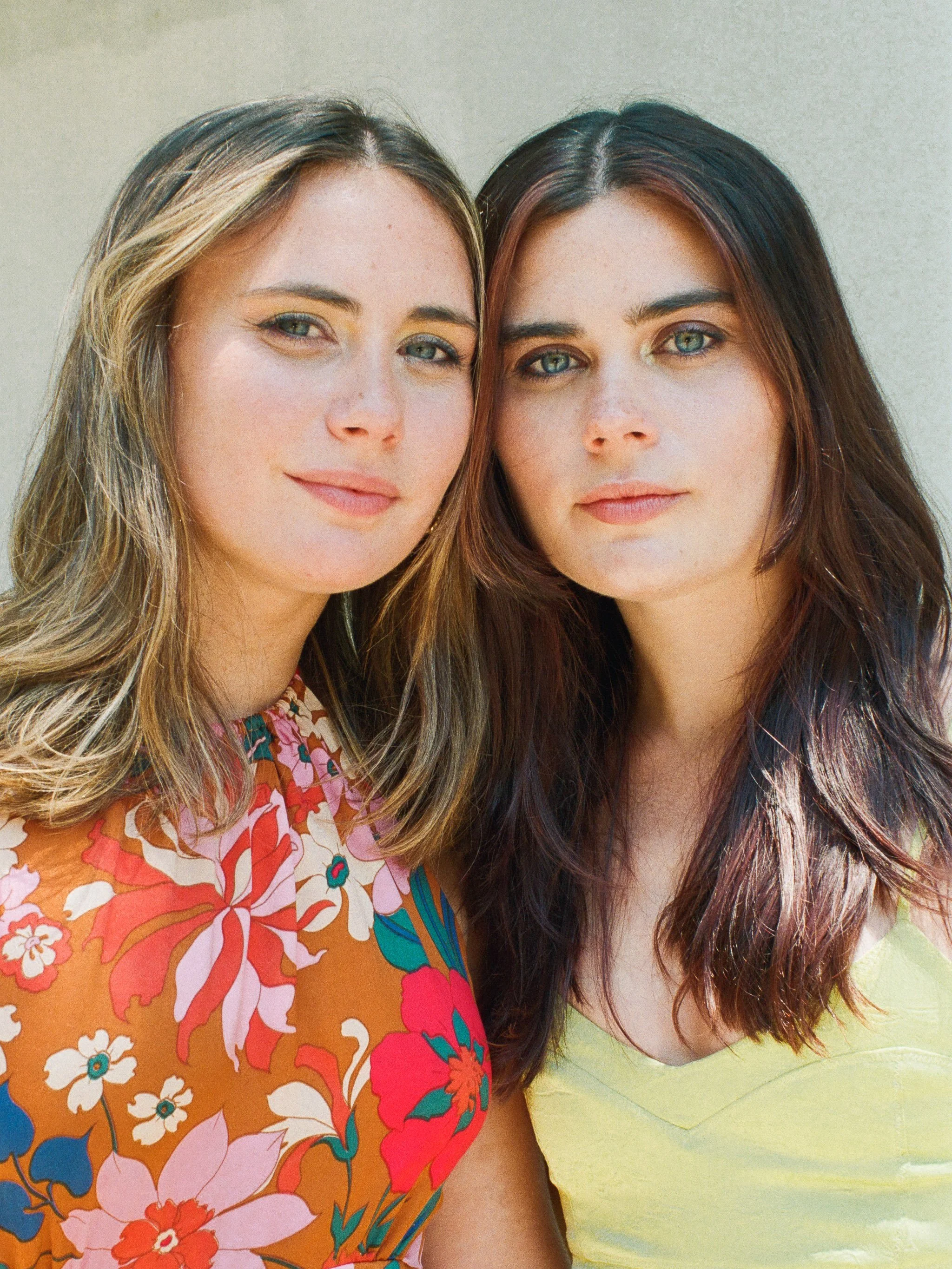 Close-up of two young women with blue eyes standing close together, one in a colorful floral dress and the other in a light yellow top, both with long brown hair and natural makeup, smiling softly at the camera.