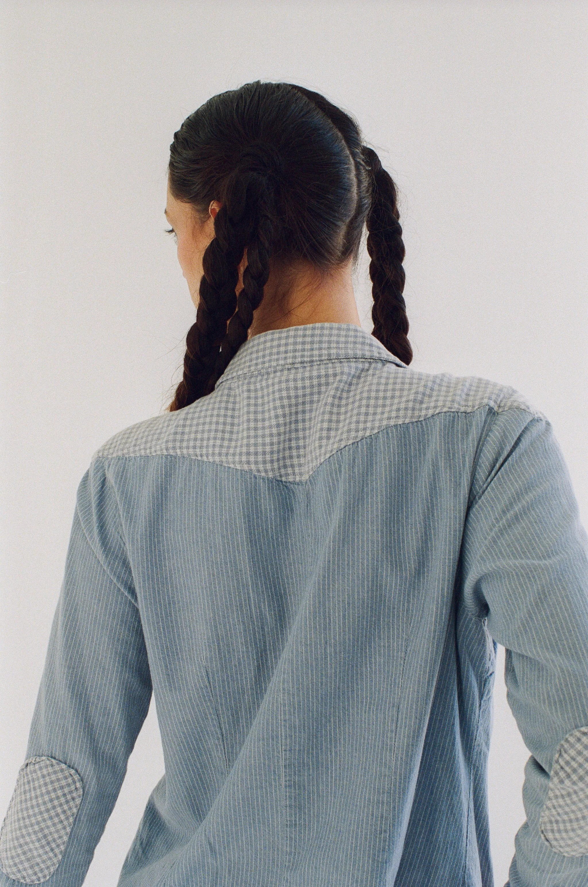 Back view of a model with dark braided hair wearing a blue and white striped shirt with checkered cuffs.