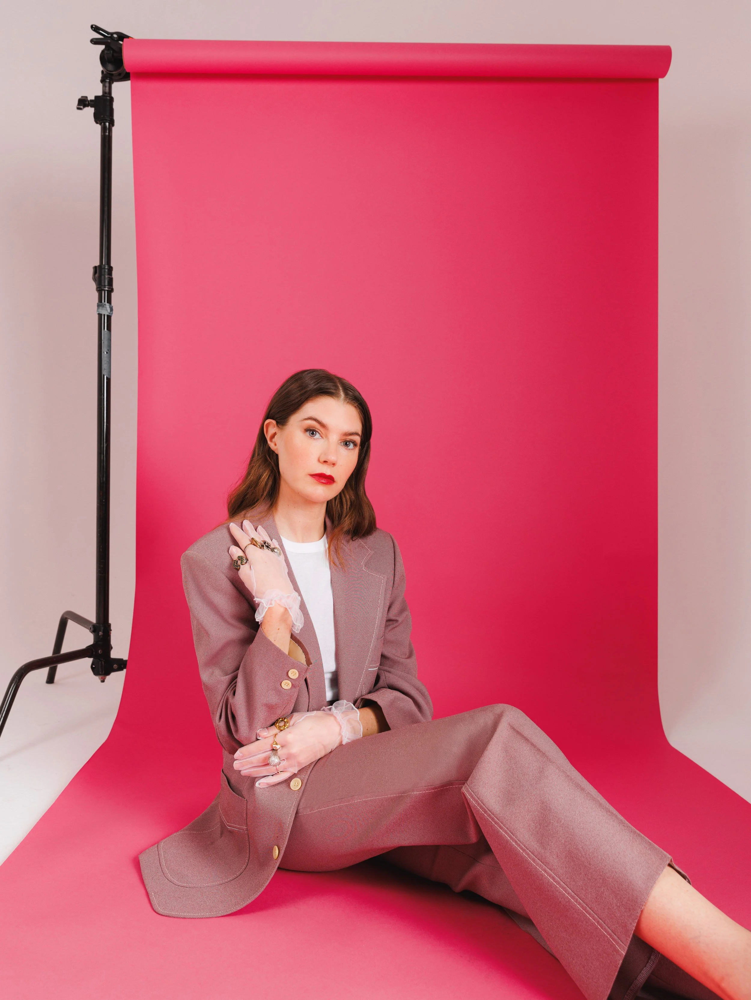 A woman with long brown hair is sitting on a pink backdrop on a visible set. She is wearing a blazer and matching pants over a white top. She is wearing several rings and delicate gloves. 