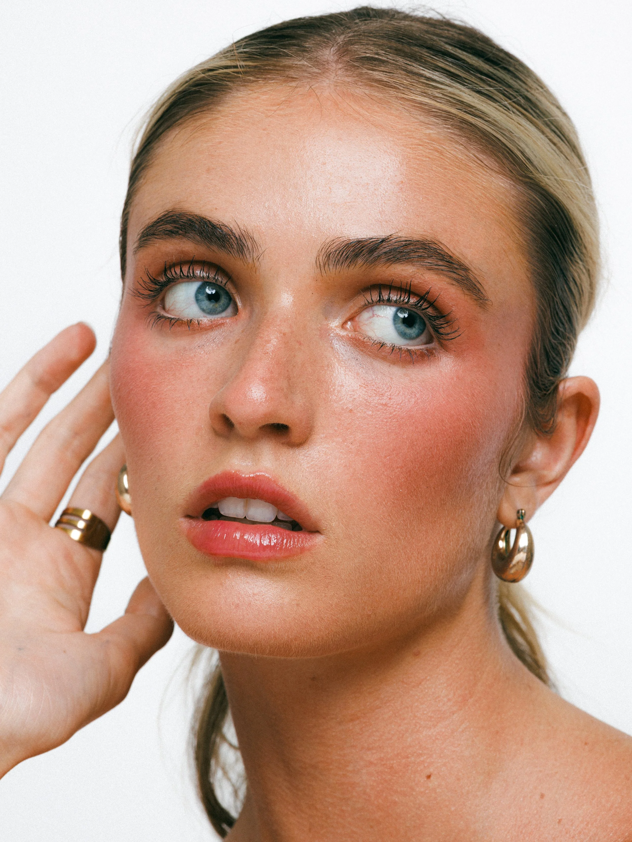 Close-up beauty portrait of a blonde model with glowing makeup, gold hoop earrings, and a sleek ponytail against a white background.