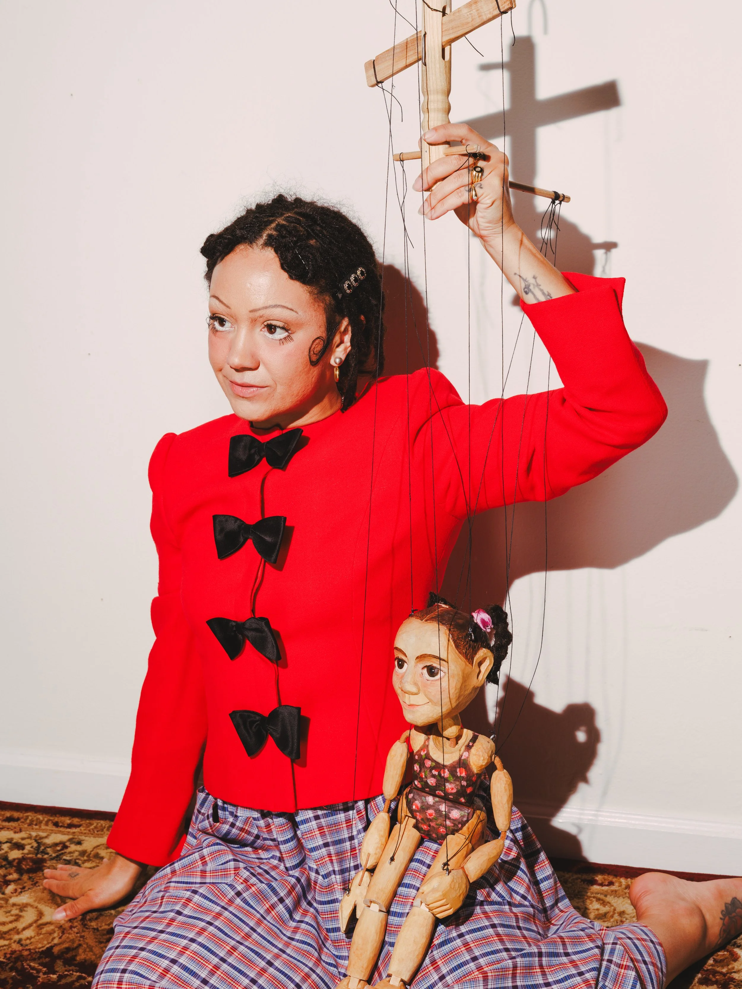 A woman dressed in a red jacket with black bows, plaid pajama pants, and a black bowtie, sitting on a carpeted floor, holding a marionette puppet with a painted face and hairstyle, in a room with a plain white wall.