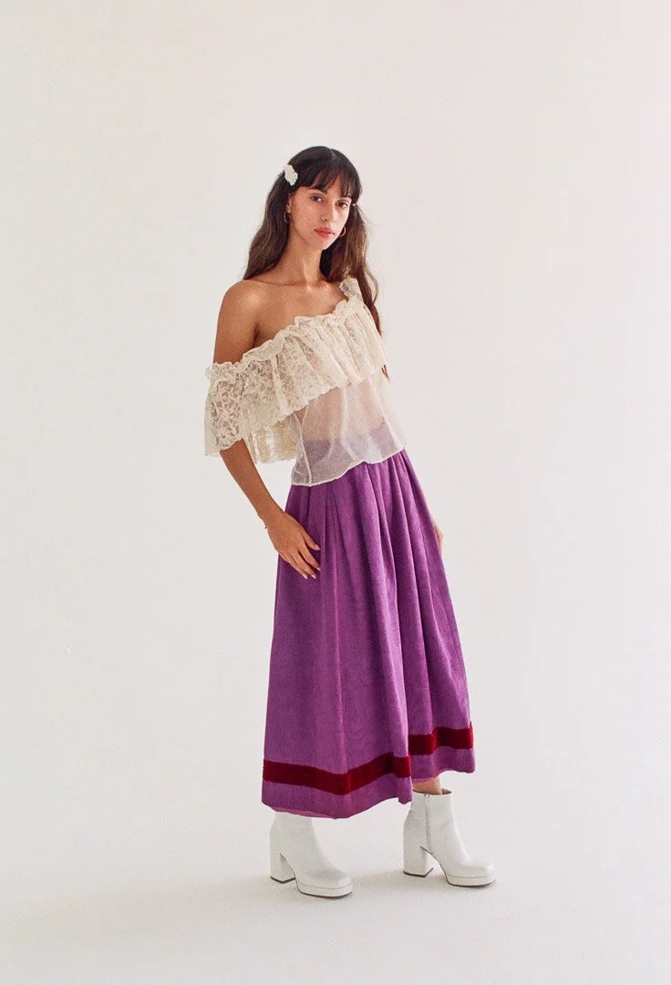 Fashion model with brown wavy hair wearing a white off-the-shoulder lace blouse, a purple pleated skirt with red bands, white ankle boots, and a hair clip, standing against a white background.