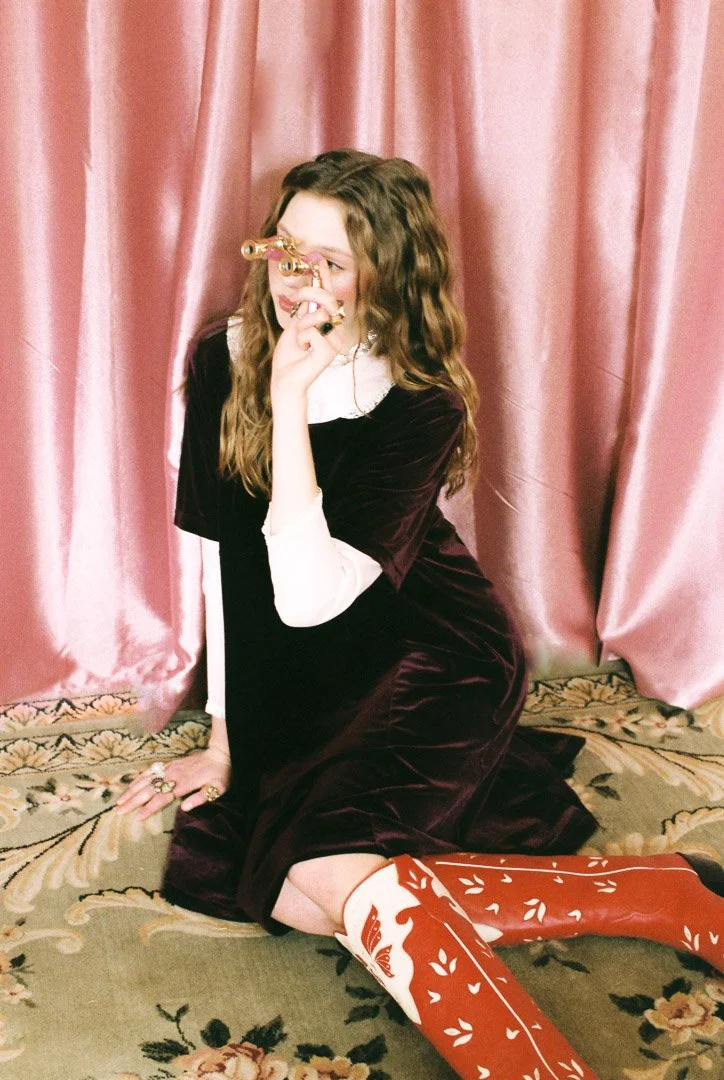 Fashion model with long wavy hair wearing a dark velvet dress with white sleeves and red cowboy boots, posing on a patterned carpet in front of pink curtains while holding a small camera to her face.