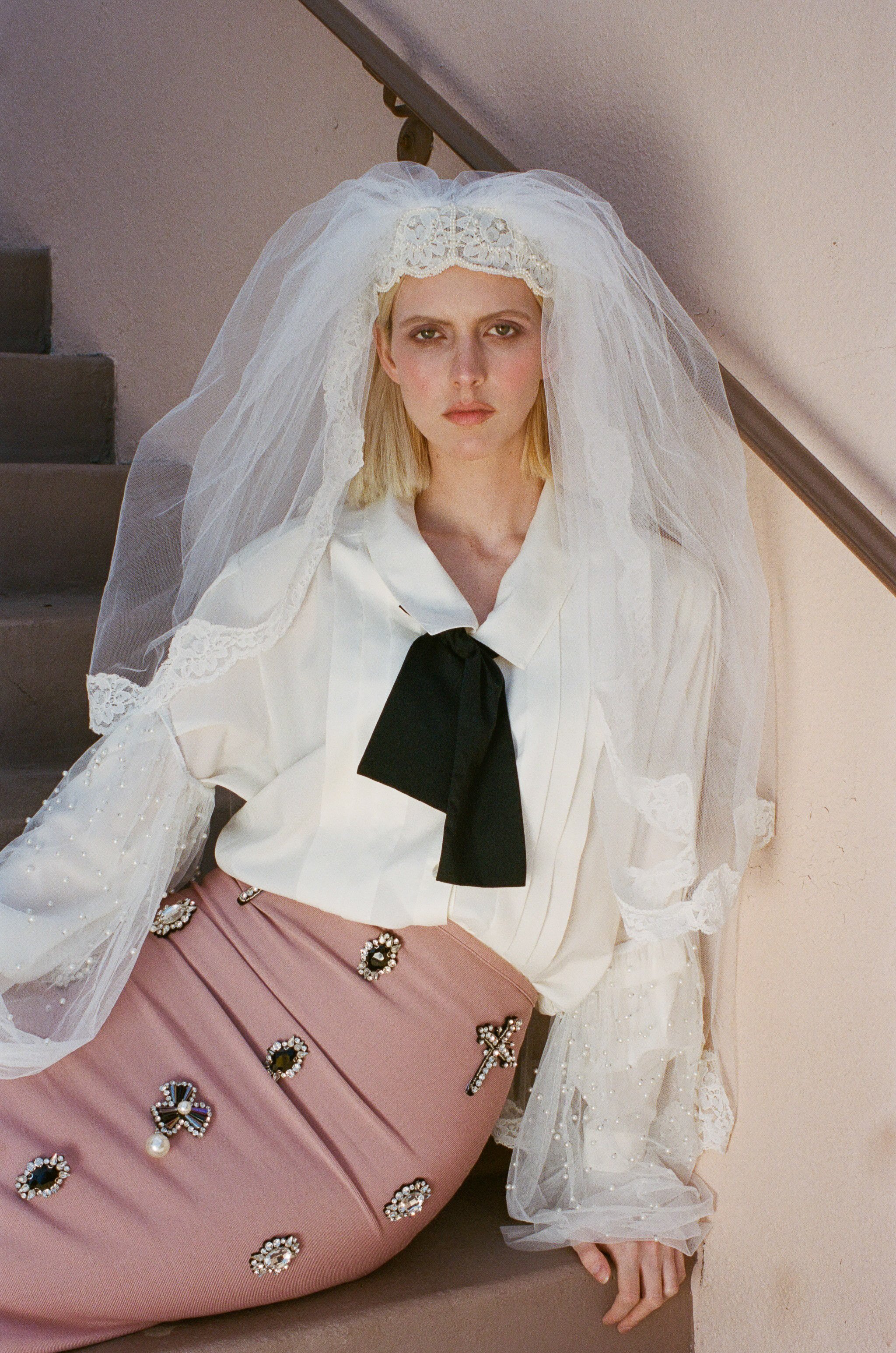Fashion model wearing a white lace veil, a white satin blouse with a black bow, and a pink skirt embellished with jeweled brooches, sitting on stairs.