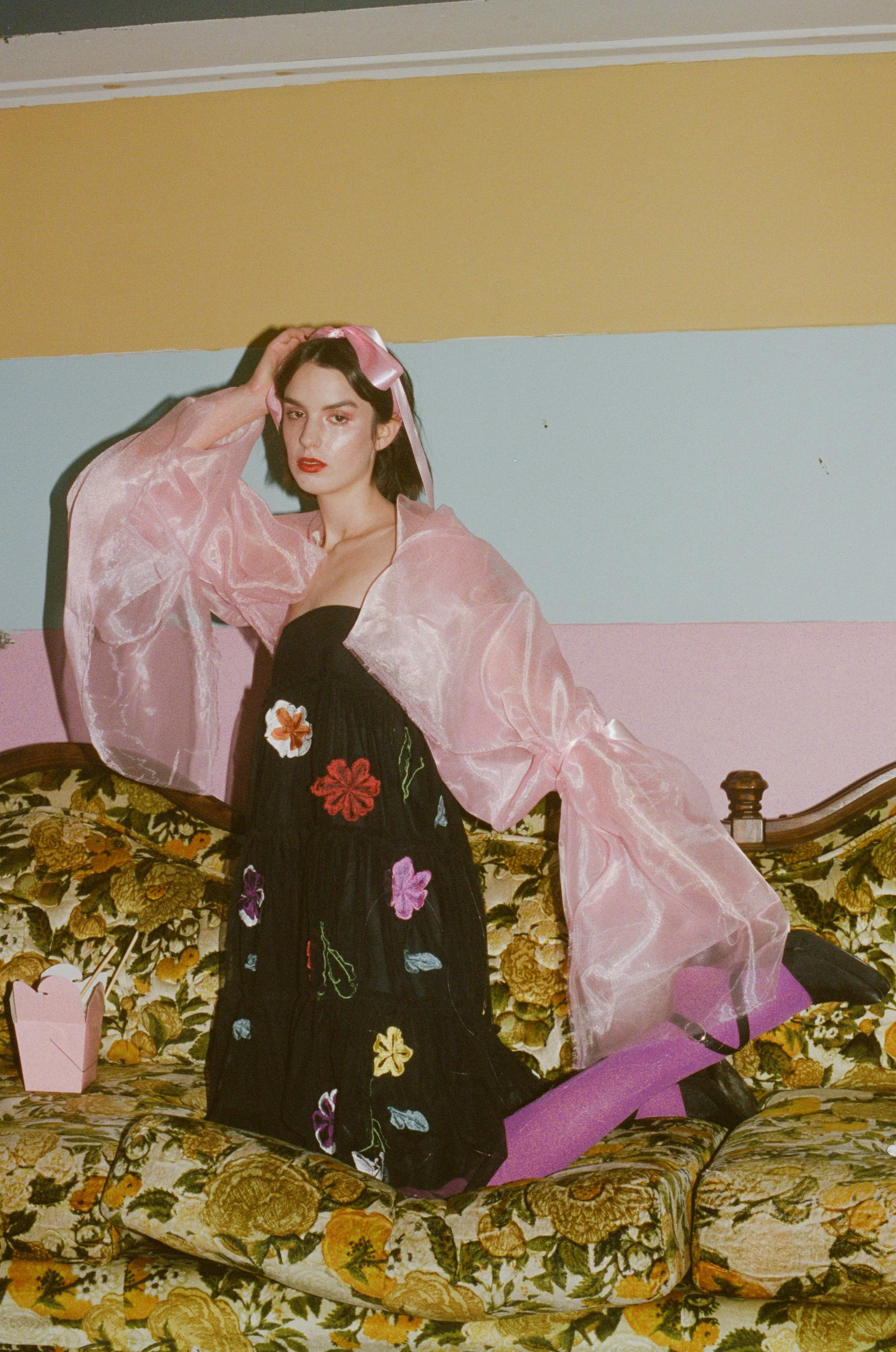 Fashion model with dark hair wearing a pink sheer jacket over a black dress with colorful floral embroidery, sitting on a vintage floral sofa against a pastel-colored wall.