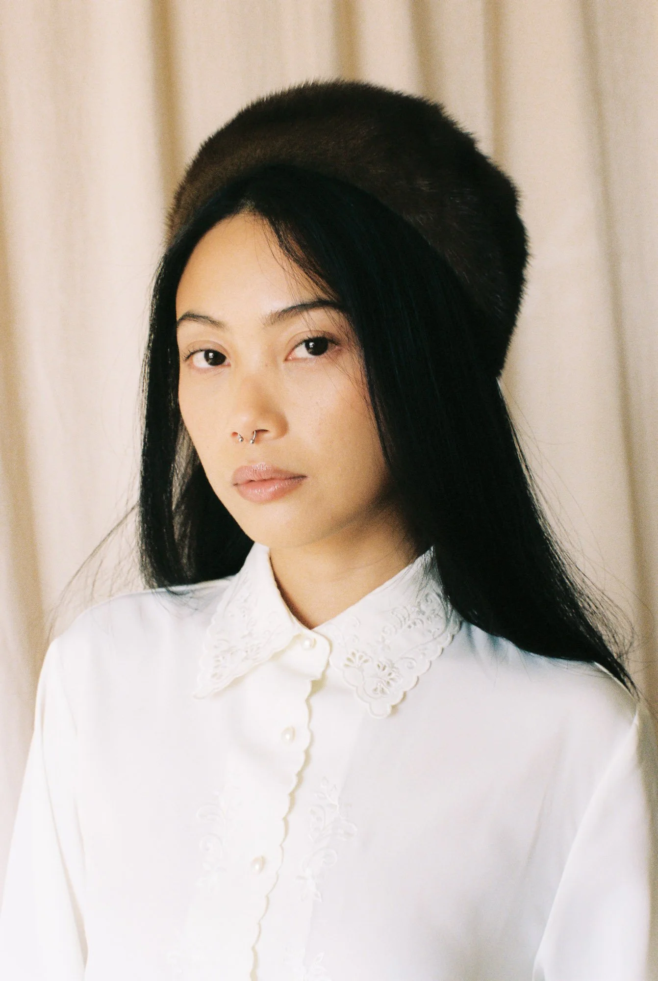 A woman with black hair, wearing a black fur hat and a white blouse with embroidered lace details.