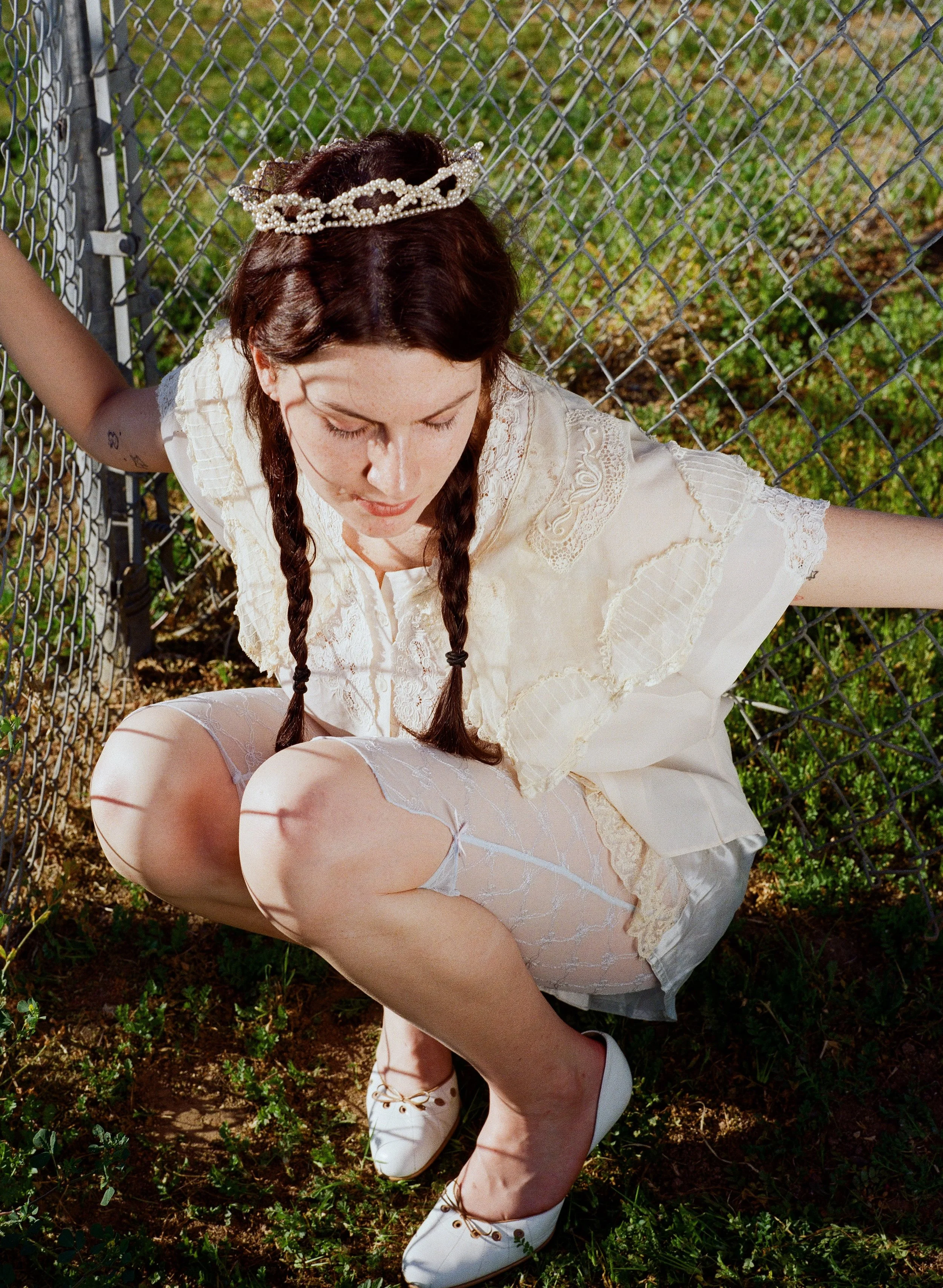 Fashion model with long braided brown hair wearing a pearl tiara, an ivory lace dress, sheer stockings, and white shoes, sitting on the ground beside a chain-link fence.