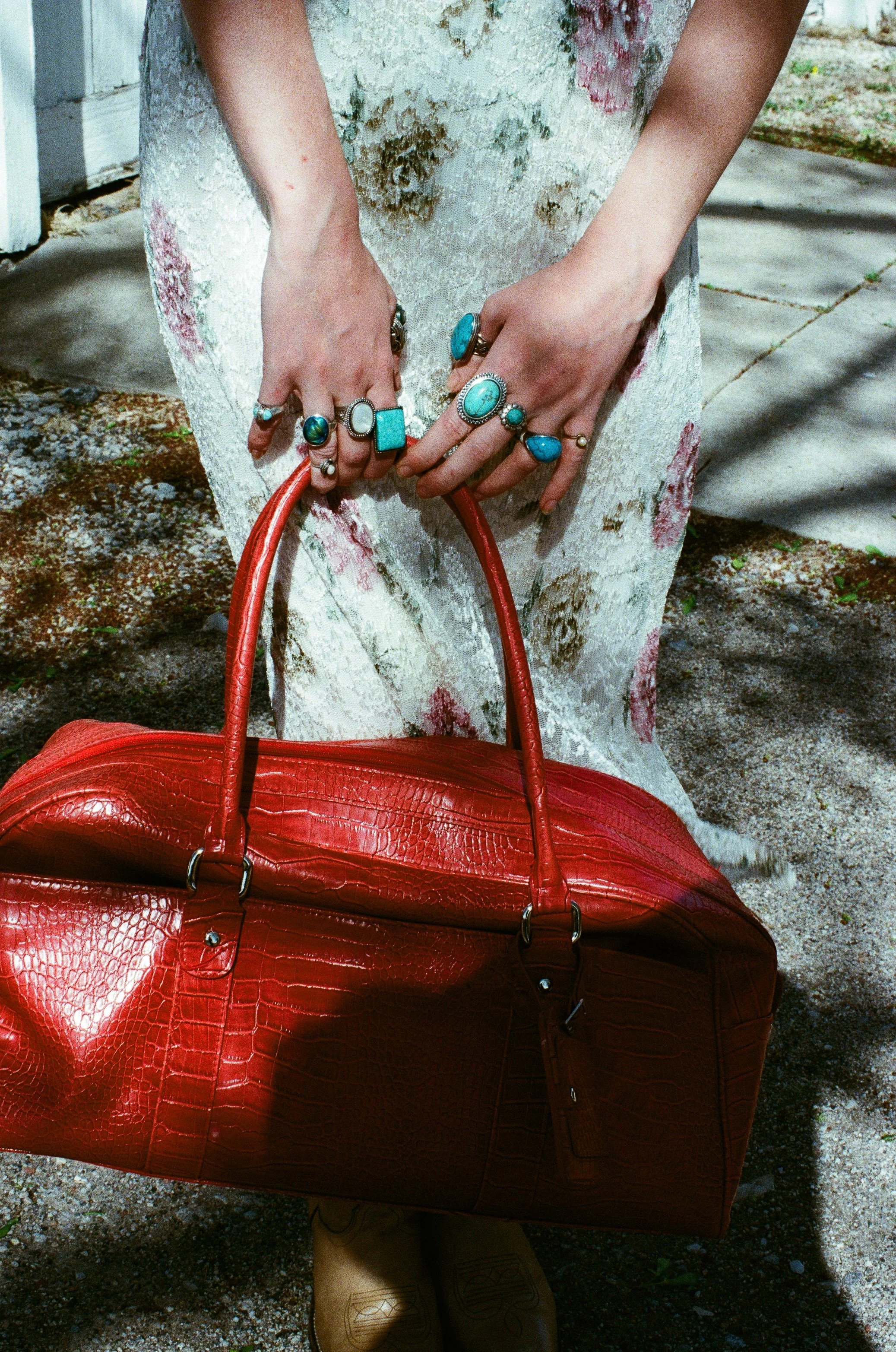 Close-up of a fashion model wearing a floral dress and rings, holding a red textured handbag outdoors on concrete pavement.