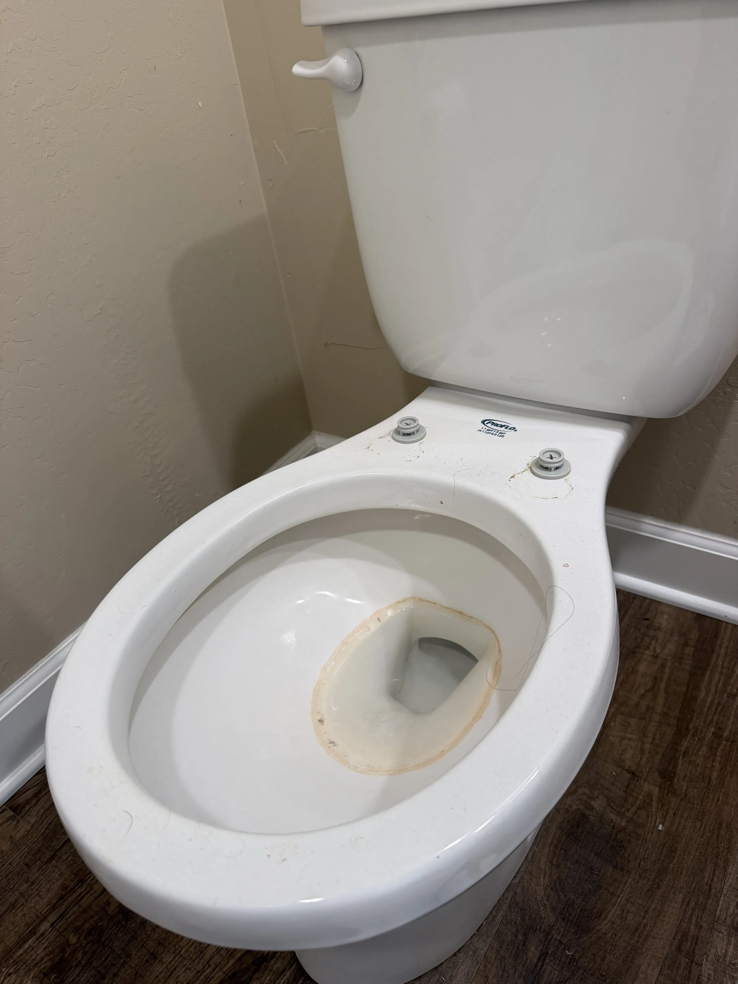 A white toilet with a stained and dirty bowl, lacking a seat, in a bathroom with beige walls and dark wooden flooring.